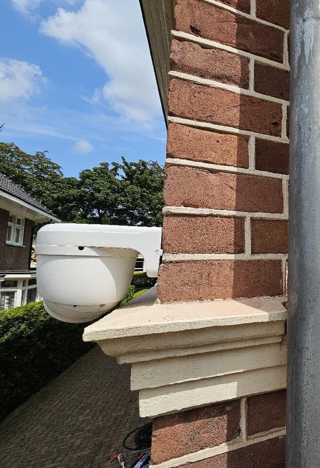 Witte dome-camera op een beugel tegen de hoek van een bakstenen pilaster, vlak boven de geprofileerde stenen lijst