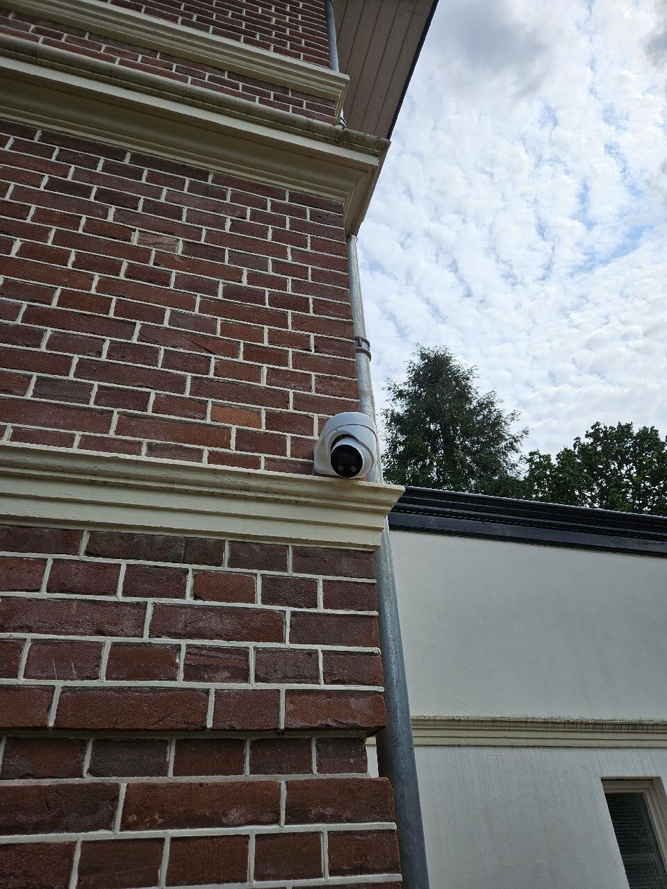 Witte dome-camera gemonteerd op een bakstenen gevel naast de regenpijp, zonder zichtbare kabel eromheen