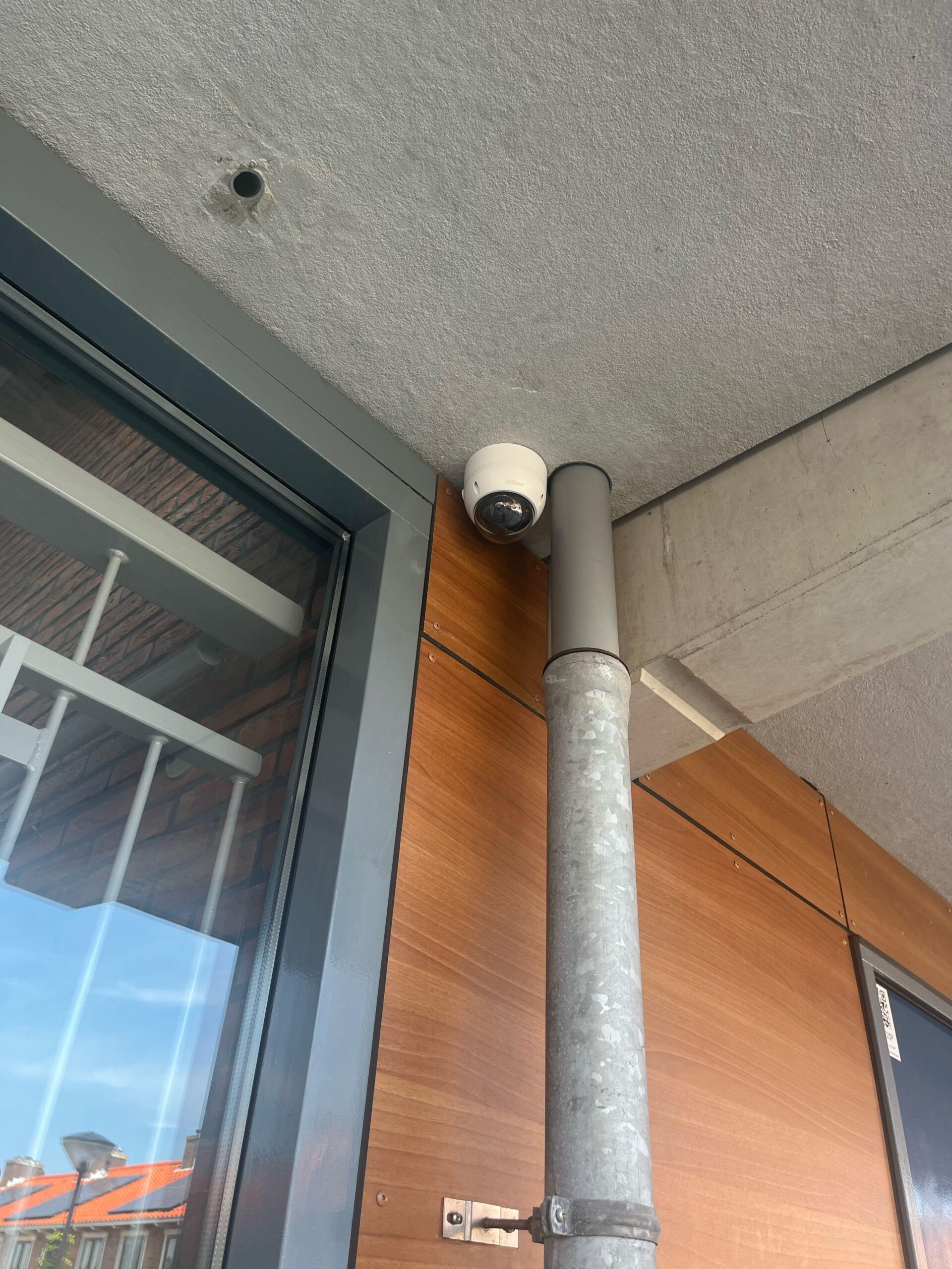 Dome-camera onder een betonnen luifel, direct naast een verzinkte regenpijp langs de houten gevelbekleding