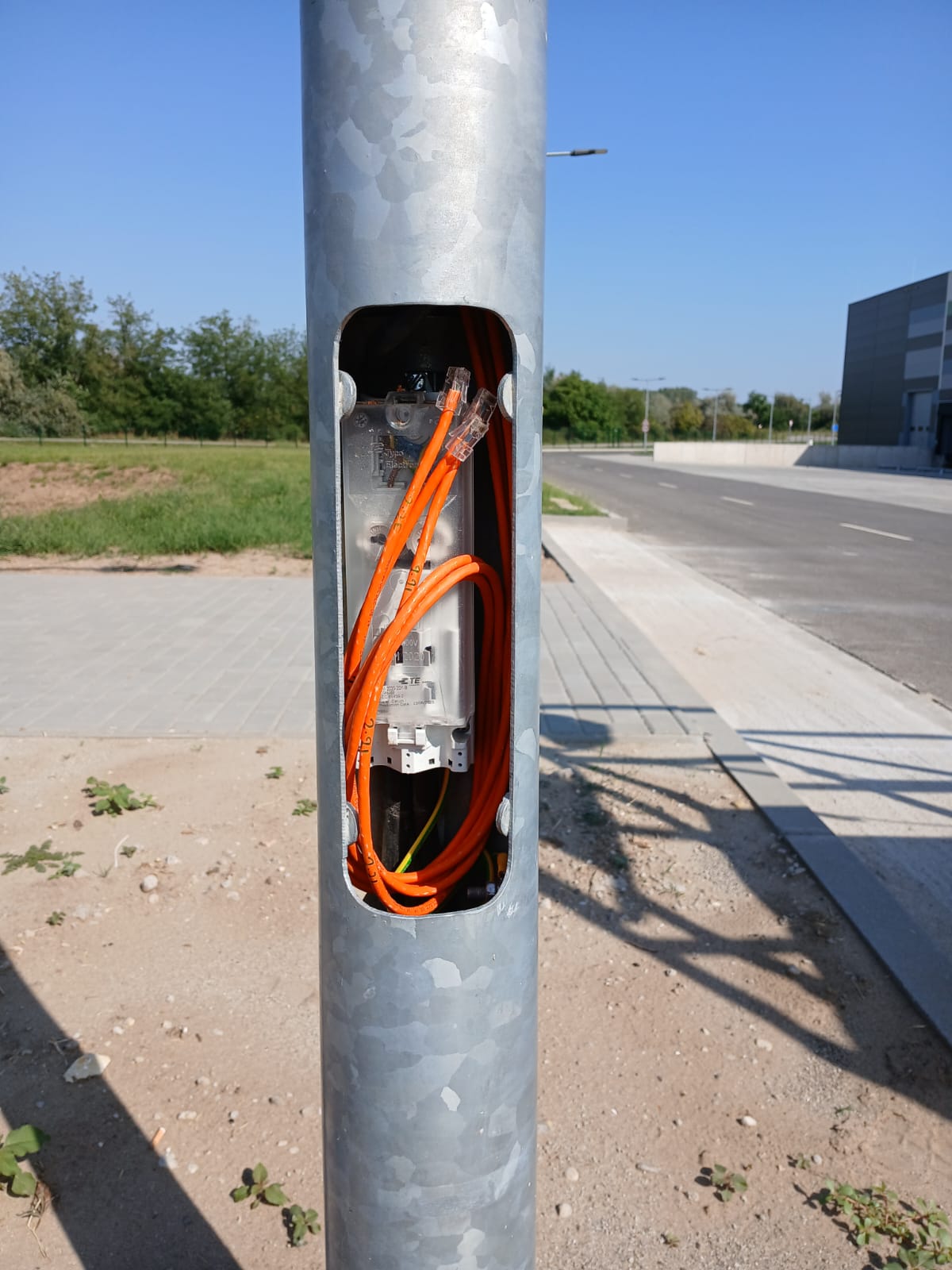 Open voet van een lichtmast met oranje netwerkkabel, RJ45-stekkers en een aansluitblok, langs een weg op een bedrijventerrein