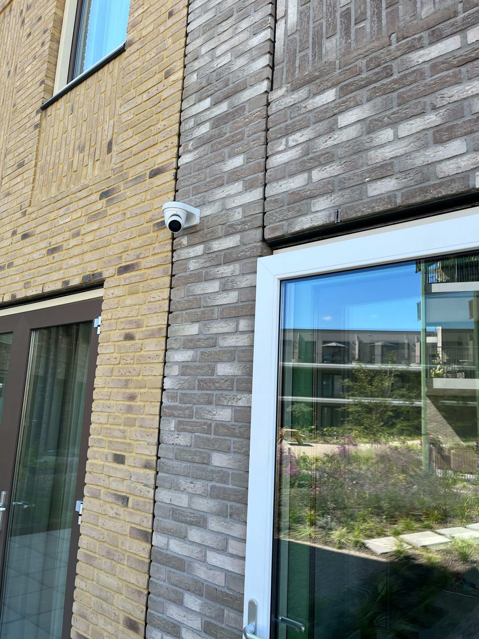 Witte dome IP-camera aan een bakstenen gevel naast een grote tuindeur, met zicht op de binnentuin in de raamweerspiegeling