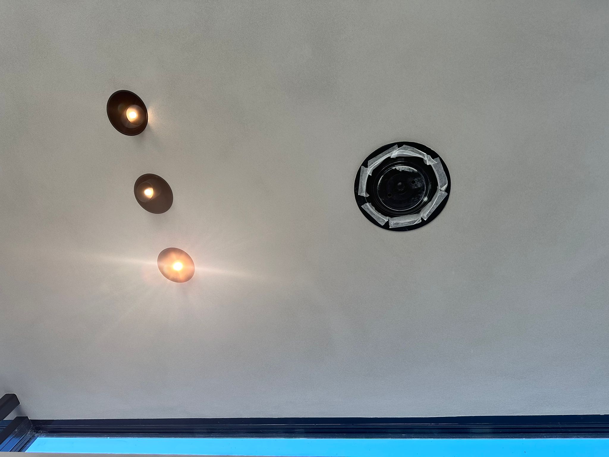 Zwarte inbouwspeaker en drie warme inbouwspots in een gestuct plafond, de speaker nog afgeplakt voor het schilderwerk