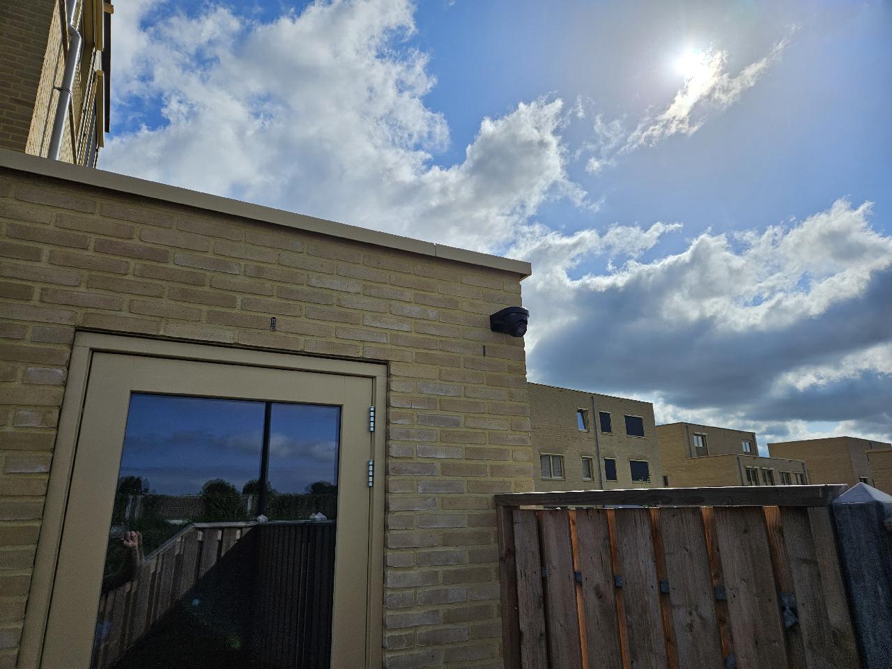Zwarte dome-camera op de hoek van een lichte bakstenen woning met tuin en schutting, blauwe lucht op de achtergrond