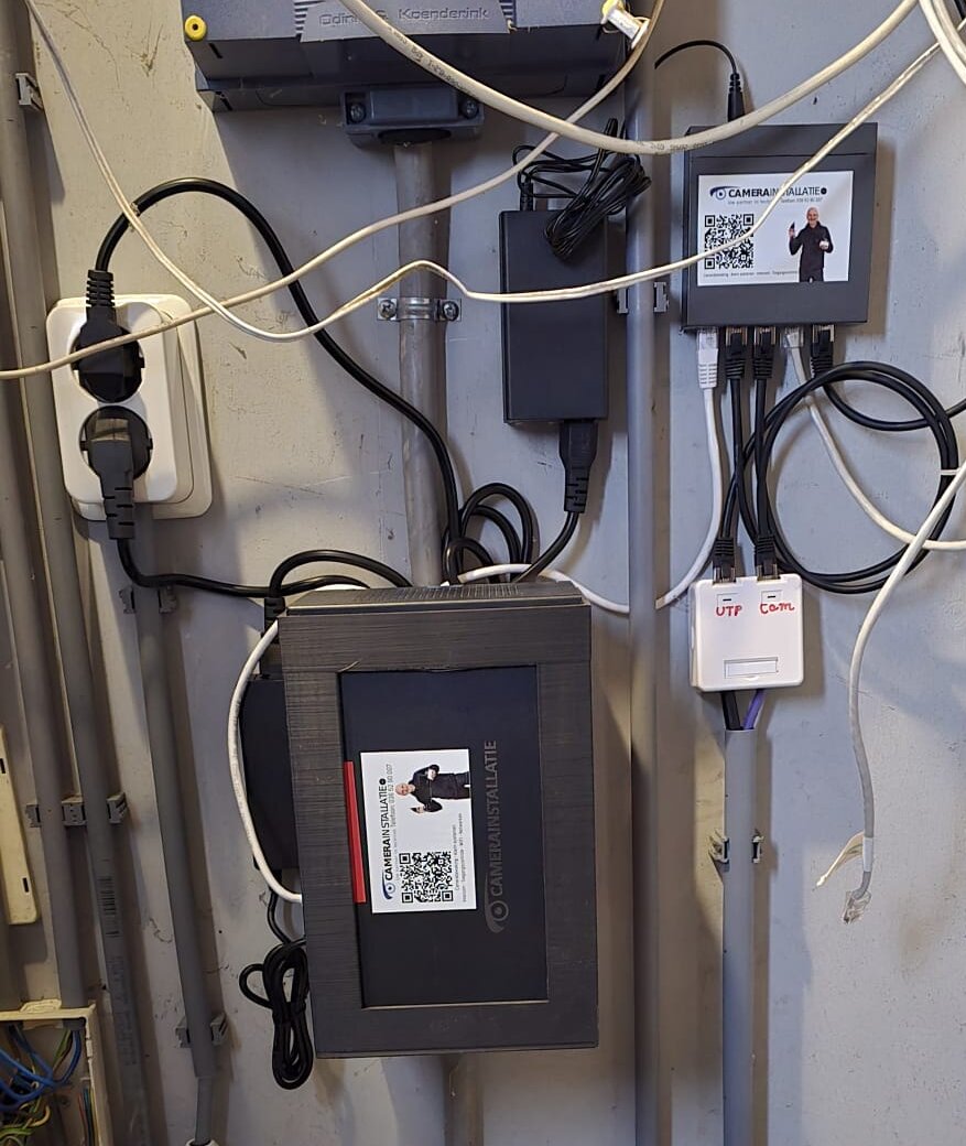 Recorder en PoE-switch met CameraInstallatie-stickers in een meterkast, met genummerde UTP- en cameraverbindingen