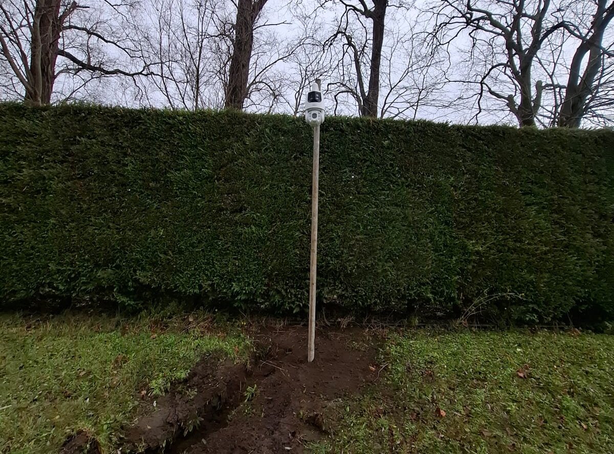 Camera met dubbele lens boven op een paal voor een hoge haag, met de verse kabelsleuf aan de voet