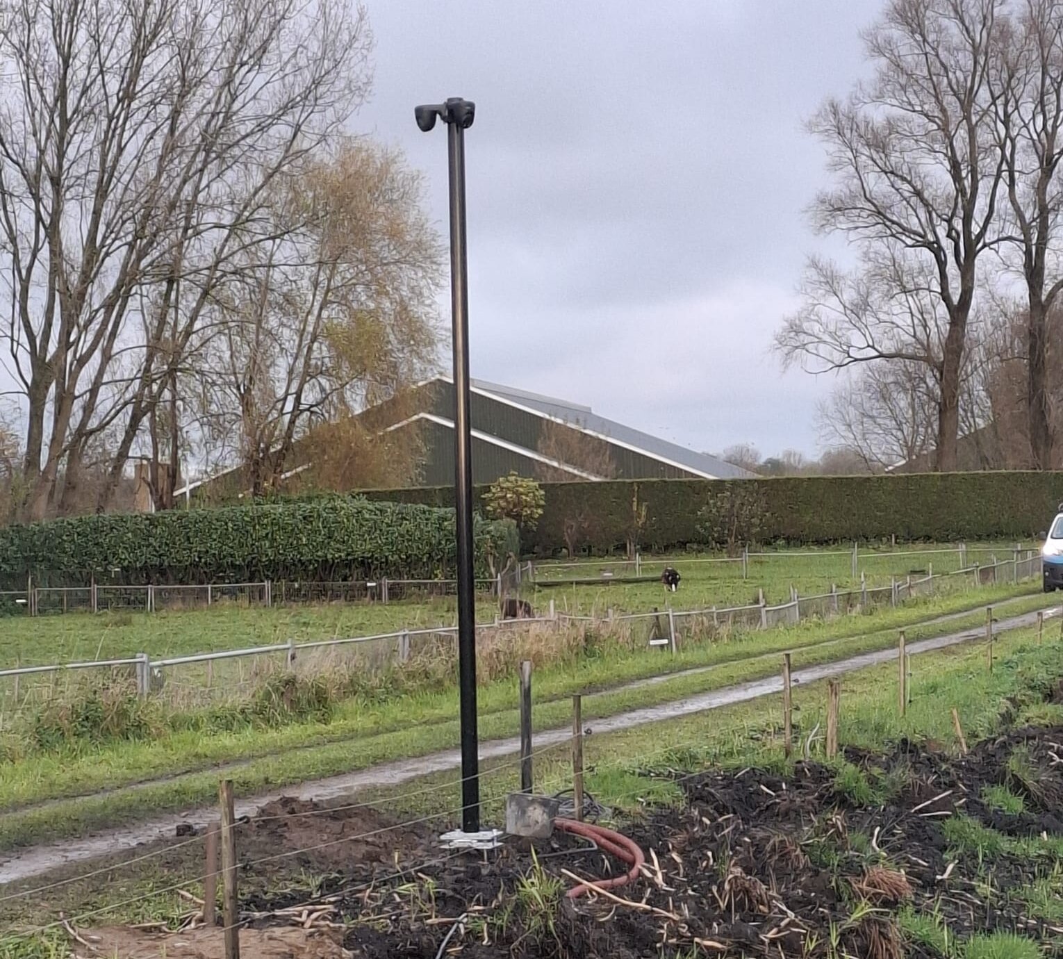 Zwarte camera-mast met dubbele lens camera bovenop, geplaatst langs een pad met heggen en een schuur op de achtergrond