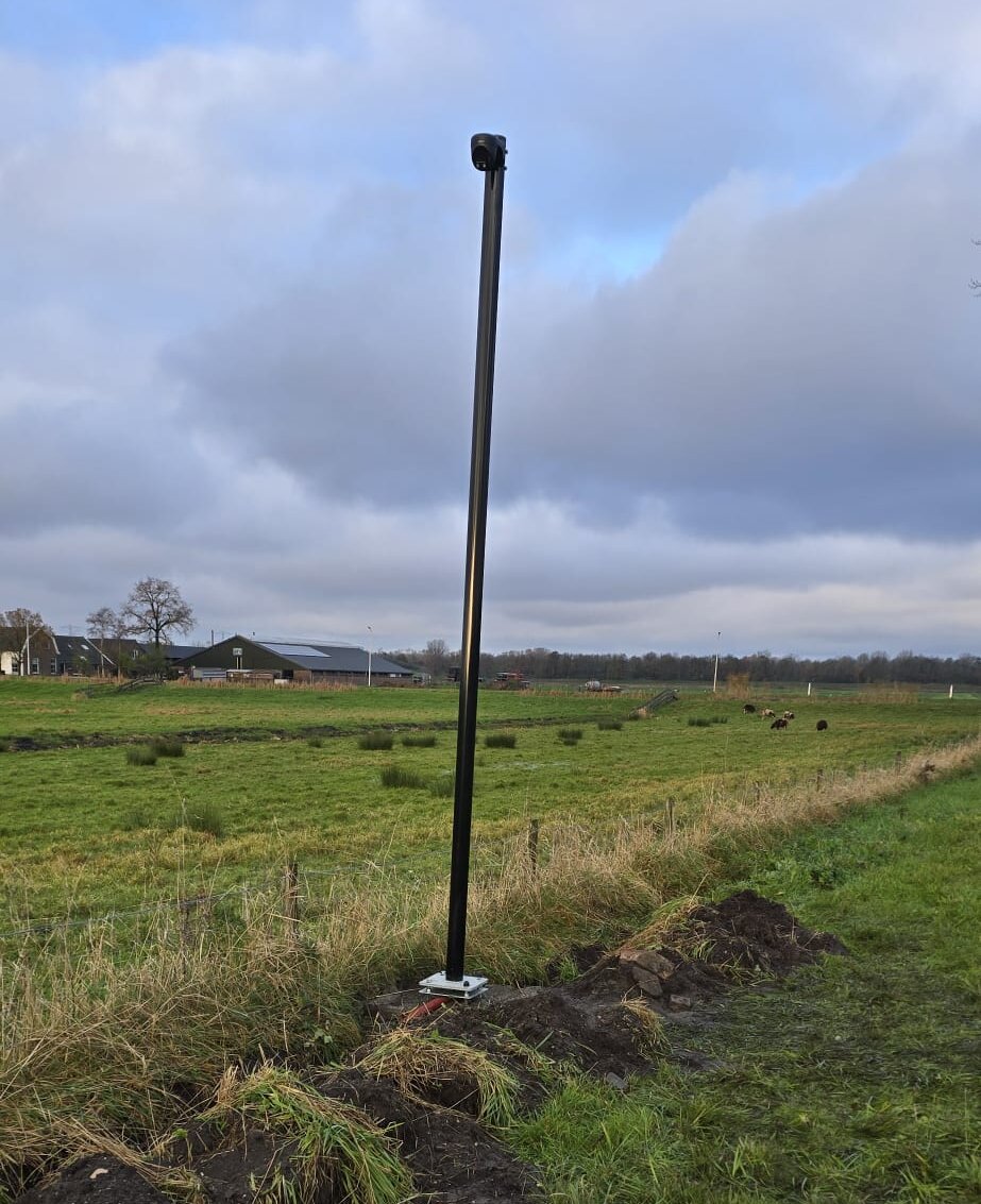 Zwarte camera-mast met camera bovenop, nieuw geplaatst in een weiland bij een woning met boerderij op de achtergrond