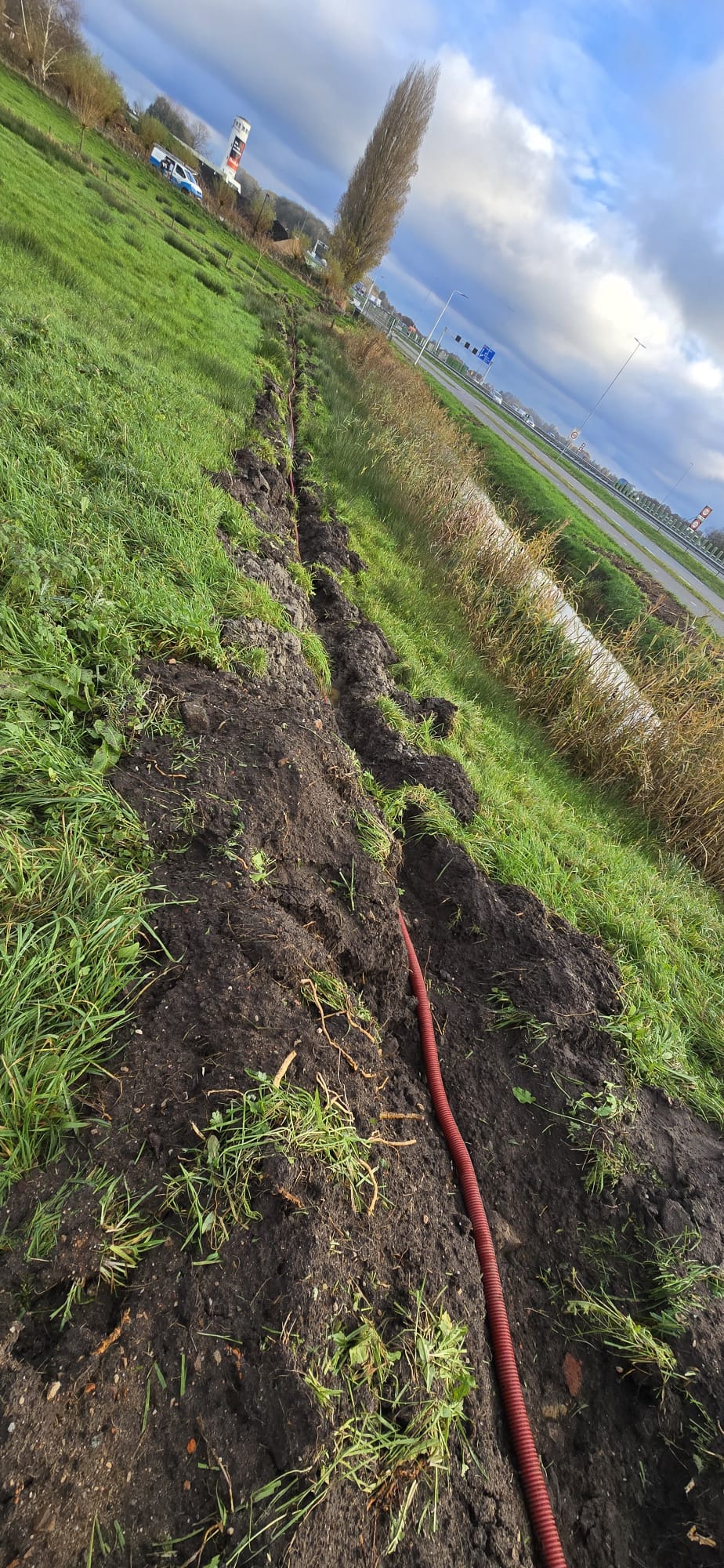 Gegraven sleuf door een grasveld met een rode mantelbuis erin voor de bekabeling naar een camera-mast