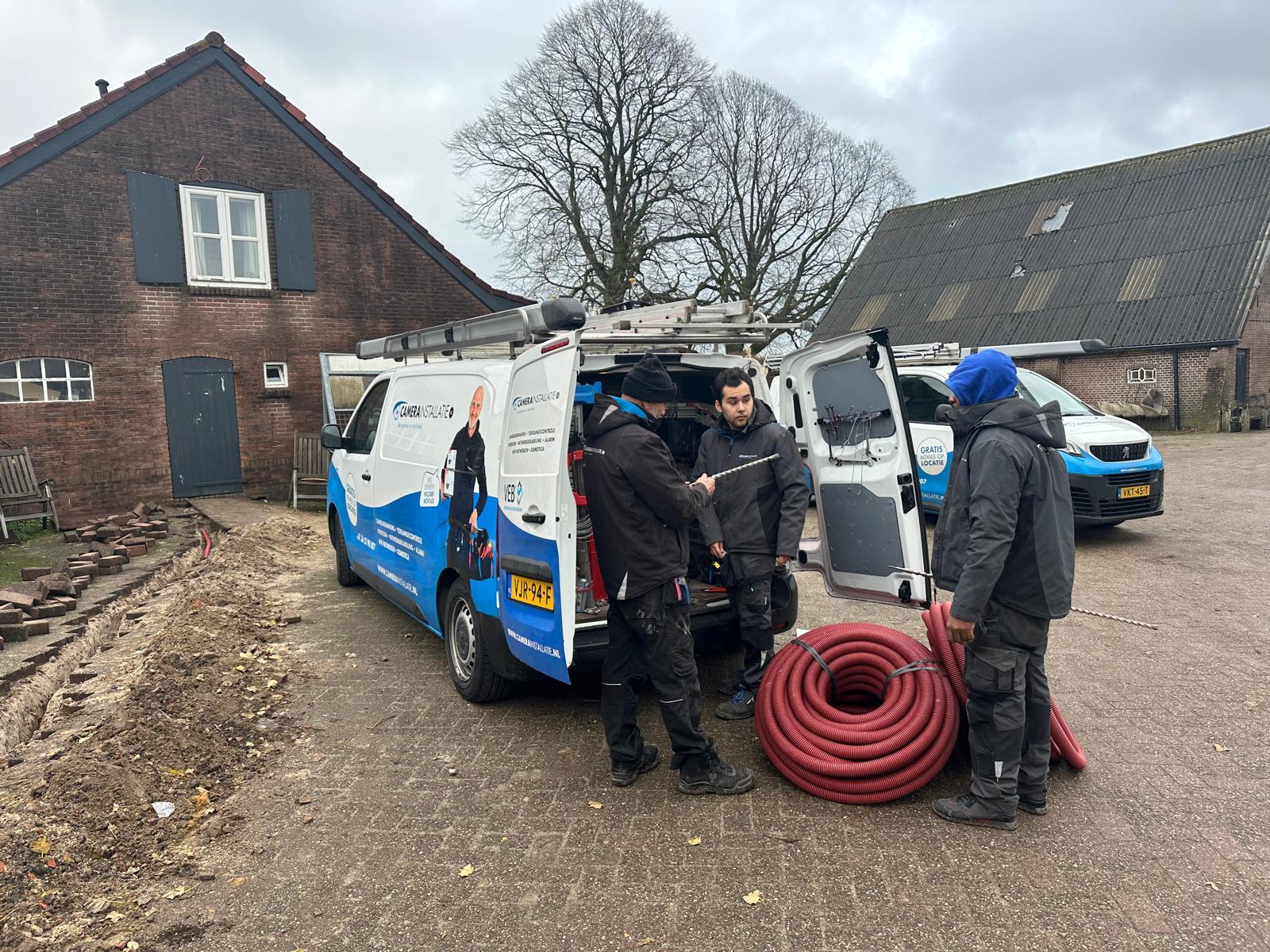 Drie monteurs van CameraInstallatie bij de herkenbare bedrijfsbus op een erf, met een rol rode kabelbuis en een gegraven sleuf