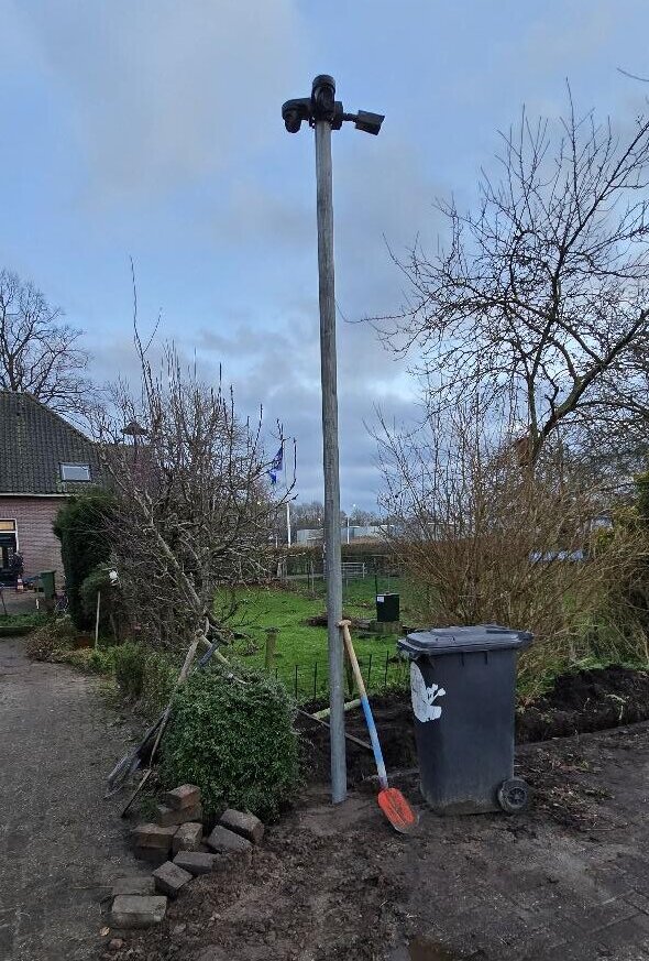 Grijze paal met twee camera’s bovenin langs het erf, met een schep en losse klinkers van het graafwerk ernaast