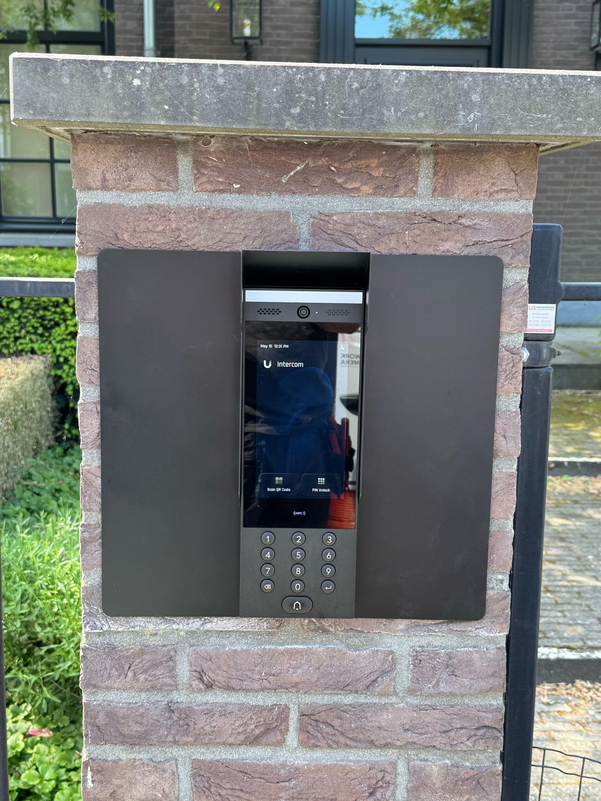 UniFi Access deurstation met touchscreen en keypad, vlak ingebouwd in een bakstenen toegangspilaar met natuurstenen deksteen