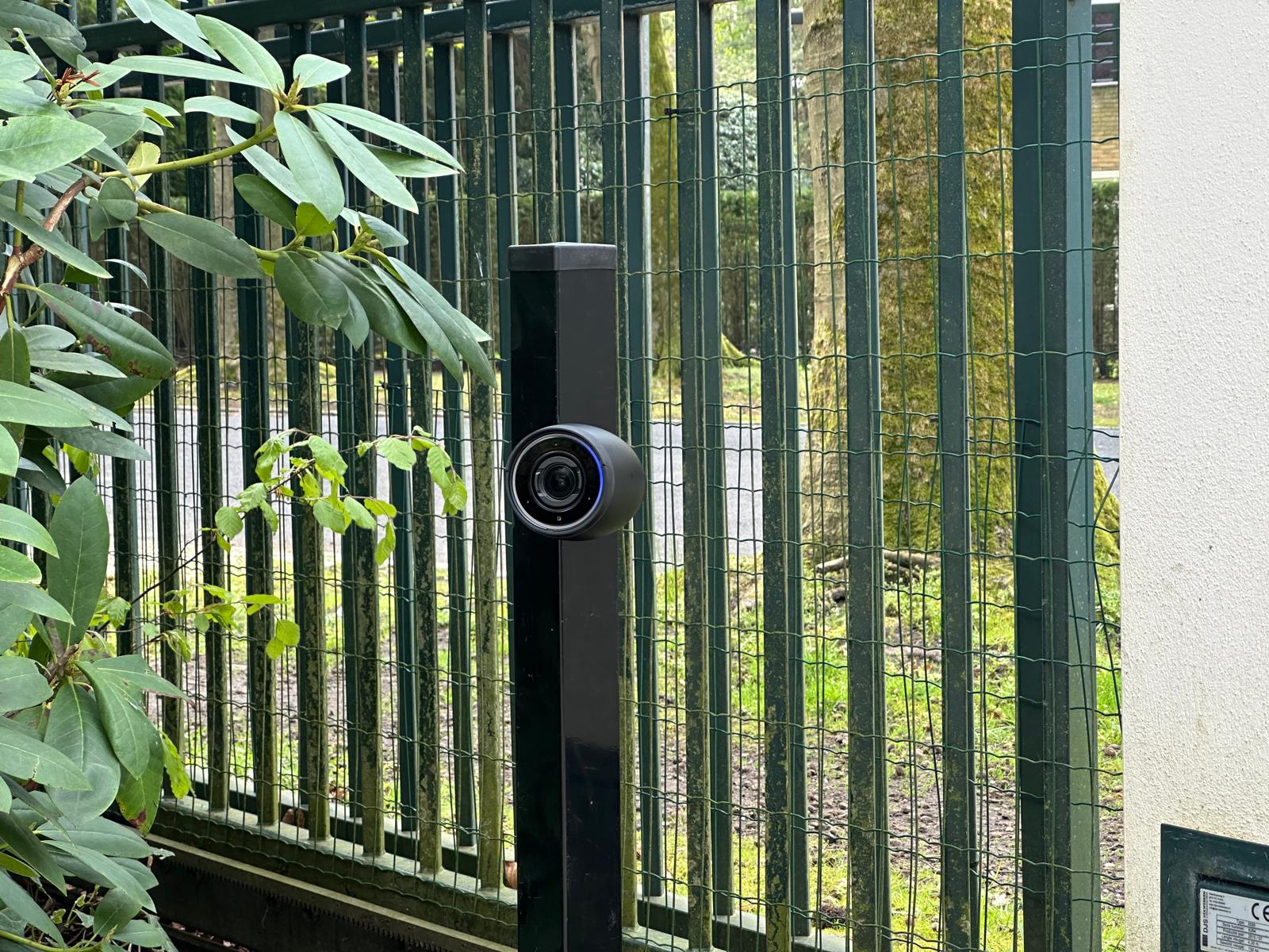 Compacte AI-camera op een zwarte paal langs een groen spijlenhek, gericht op de toegang