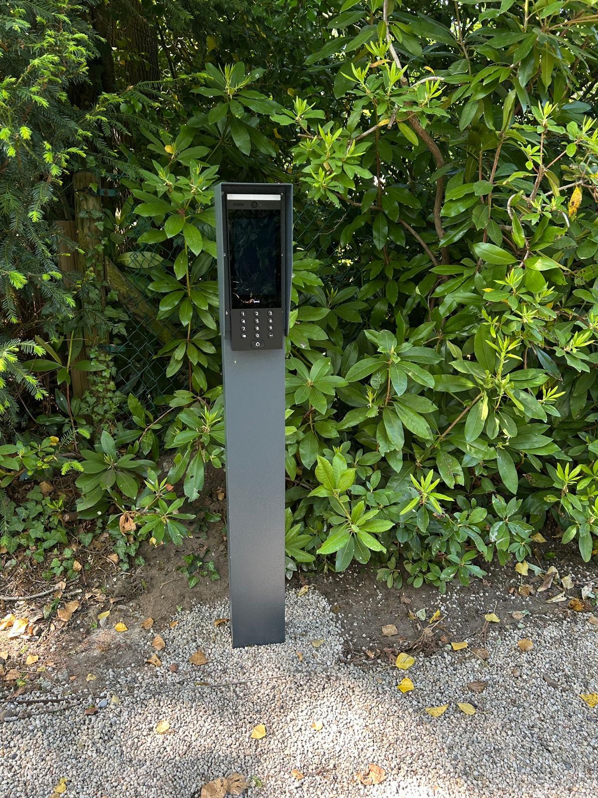 Video-intercom in een donkere toegangszuil bij een haag, met scherm en keypad