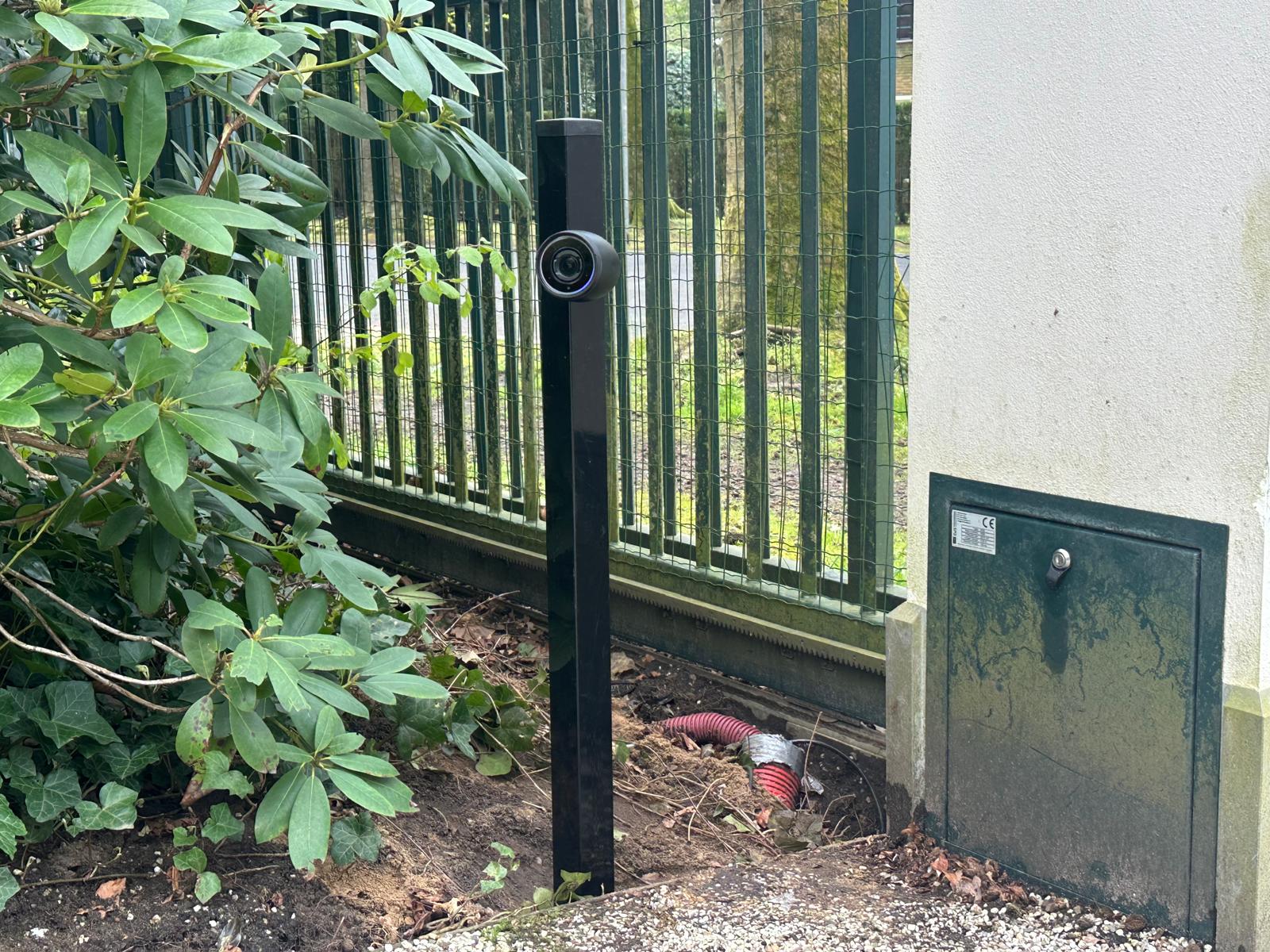 Compacte zwarte AI-camera op een lage paal naast een groen toegangshek, gericht op de oprit voor kentekenherkenning