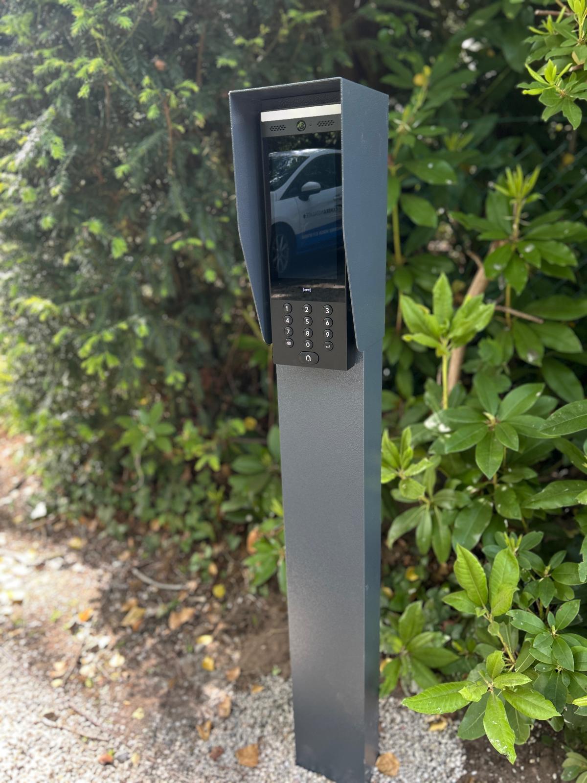 Video-intercom in een grijze toegangszuil met regenkap, het scherm spiegelt een bus van CameraInstallatie