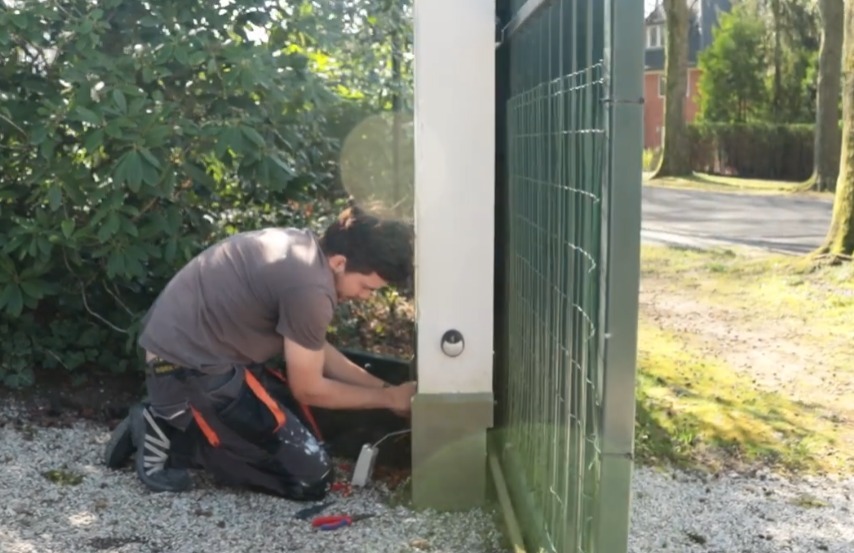 Monteur van CameraInstallatie legt geknield de bekabeling aan bij de voet van een groen toegangshek