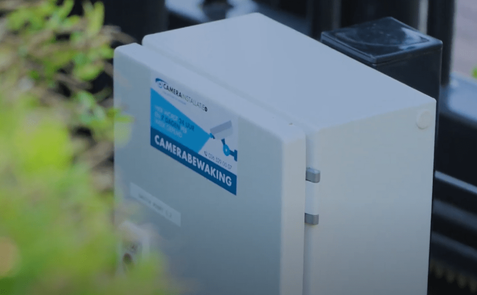 Witte buitenkast met CameraInstallatie-sticker camerabewaking, gemonteerd langs een pad op het terrein