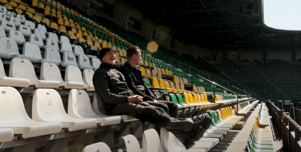 Twee teamleden van CameraInstallatie in de lege groen-gele tribune van het ADO Den Haag stadion, met zonlicht over de stoelen