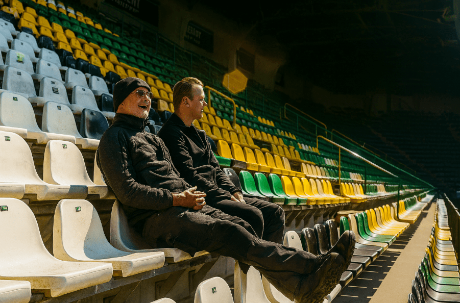 Twee mannen van het team van CameraInstallatie zitten in de groen-gele tribunestoelen van het ADO Den Haag stadion