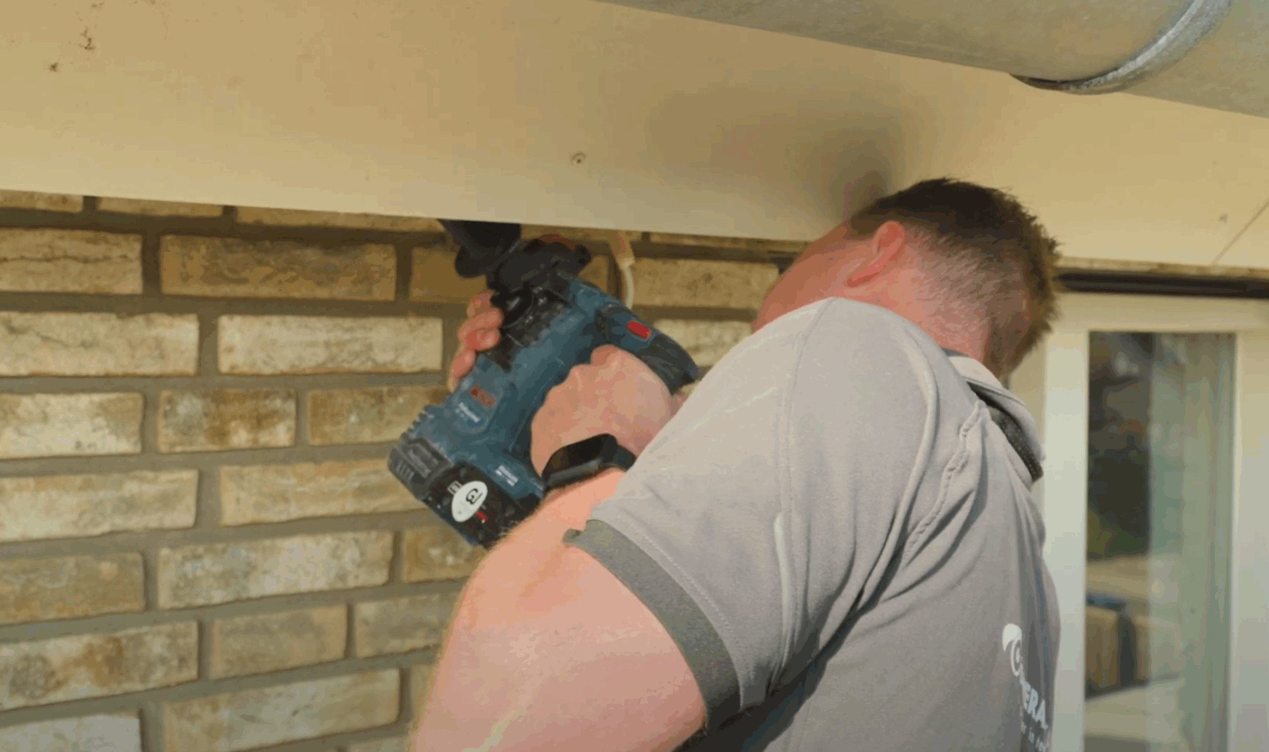 Monteur van CameraInstallatie boort met een accuboormachine onder een dakrand tijdens het aanleggen van de camerabekabeling