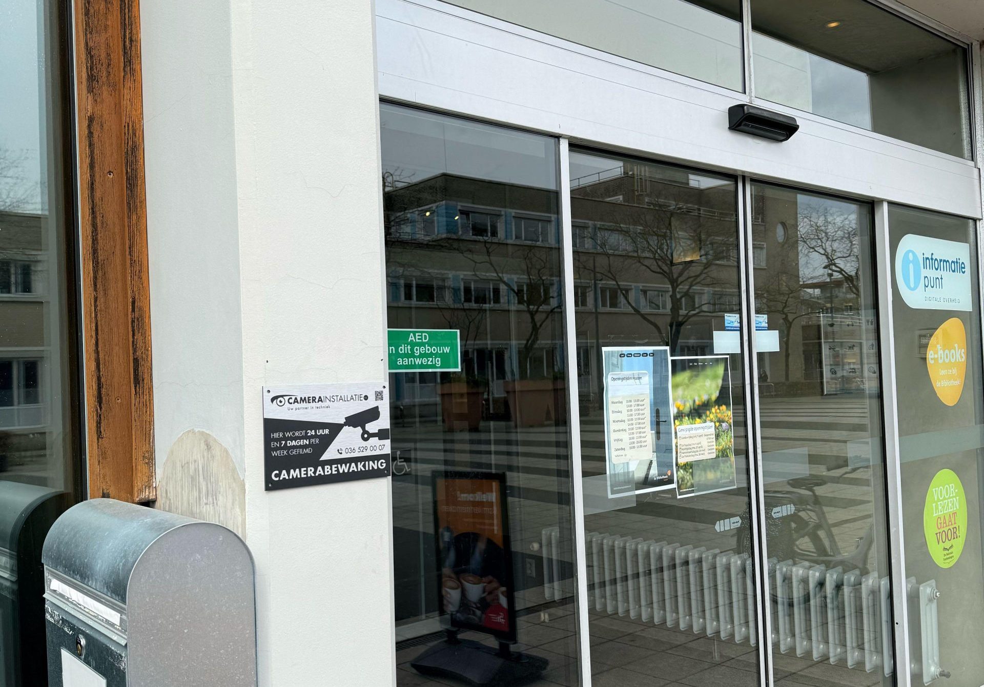 Gevel van een publiek gebouw met een CameraInstallatie.nl-bordje 'hier wordt 24 uur gefilmd' naast de schuifdeur bij de entree