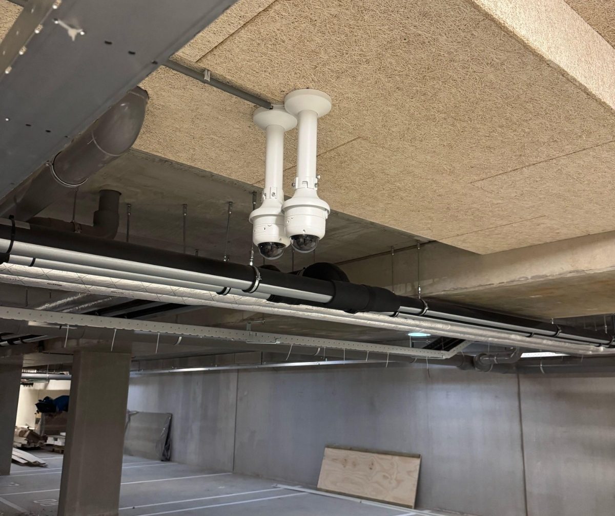 Twee witte turretcamera’s op verlengde muurbeugels onder het plafond van de parkeergarage, tussen de leidingen en luchtkanalen