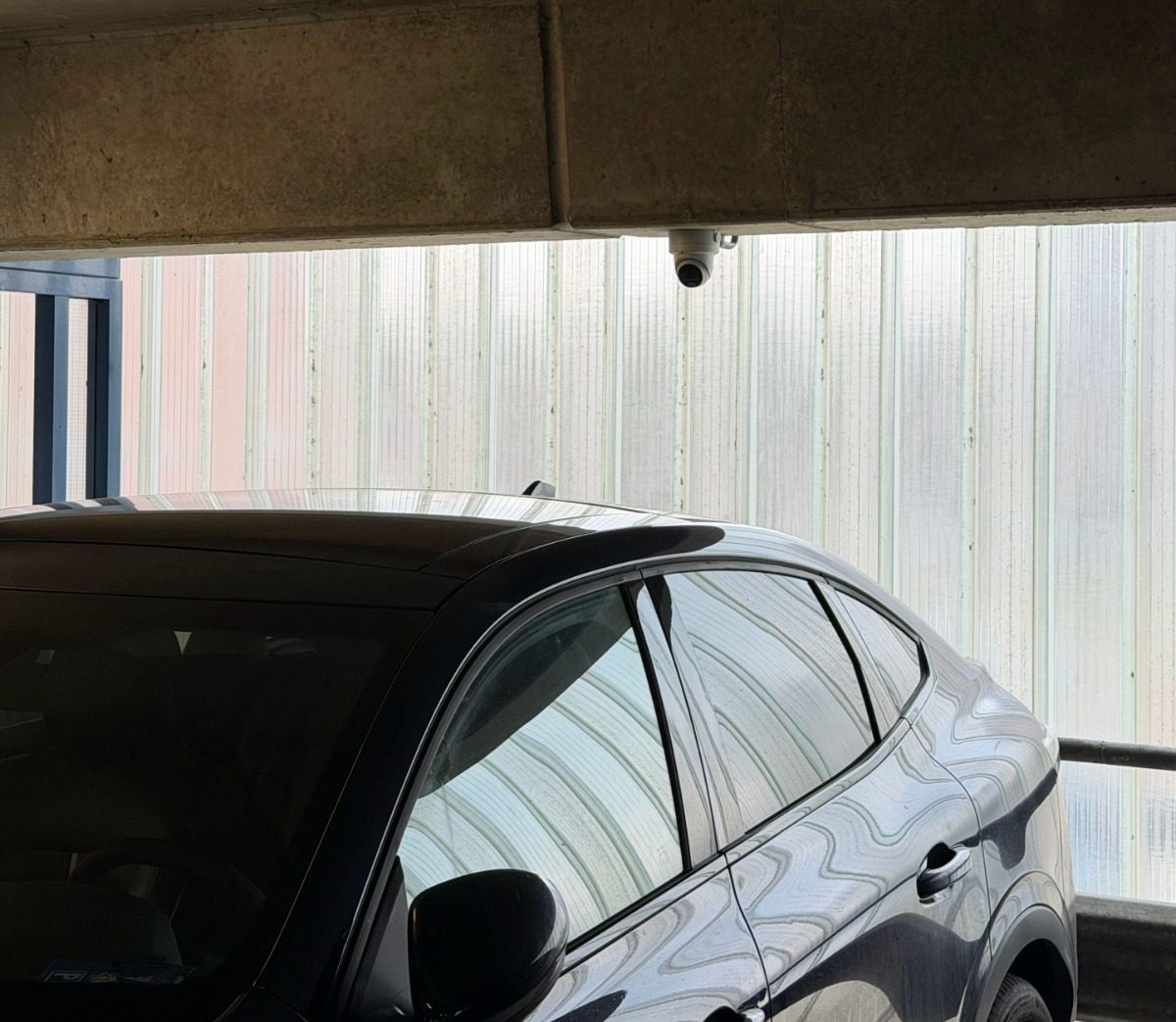 Donkere auto in de parkeergarage met een dome-camera onder de betonnen rand, tegen een lichtdoorlatende gevel