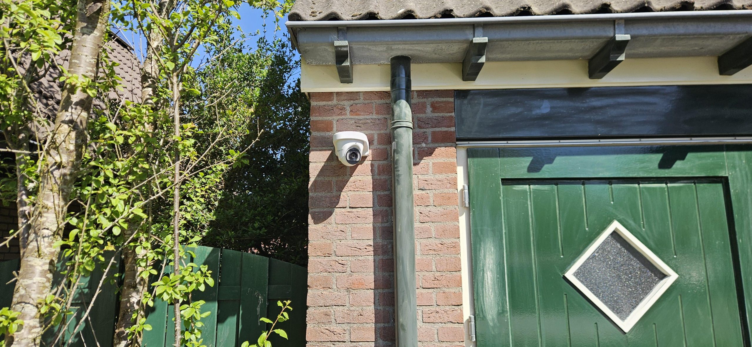 Witte Ajax dome-camera gemonteerd op een bakstenen gevel naast een groene schuurdeur, in de buitenlucht