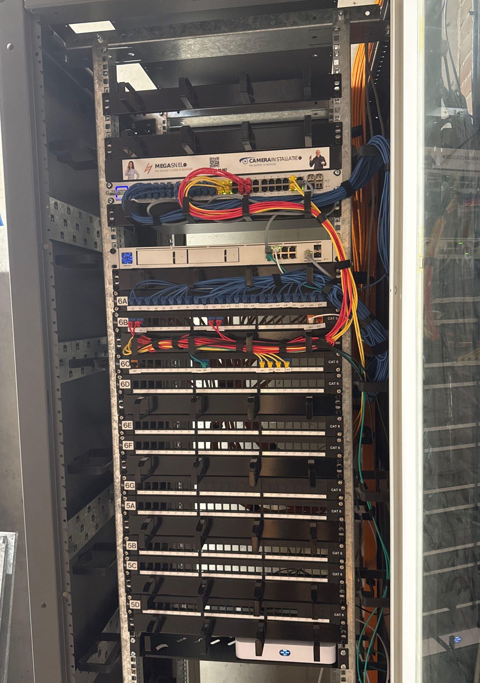 Volledige patchkast met meerdere CAT6-patchpanelen, gekleurde patchkabels en een labelstrip van CameraInstallatie en MegaSnel