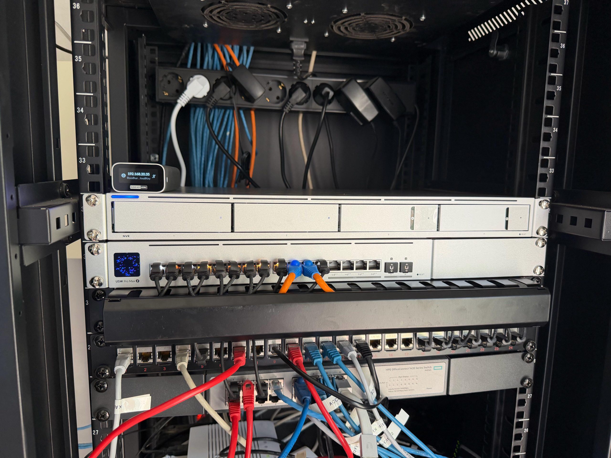 Netwerkkast met UniFi switch, netwerkvideorecorder en Cloud Key naast een patchpaneel met gekleurde patchkabels