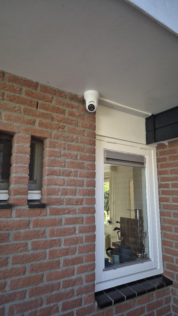 Witte Ajax dome-camera onder een overstek naast een raam van de woning, gericht op de gevel