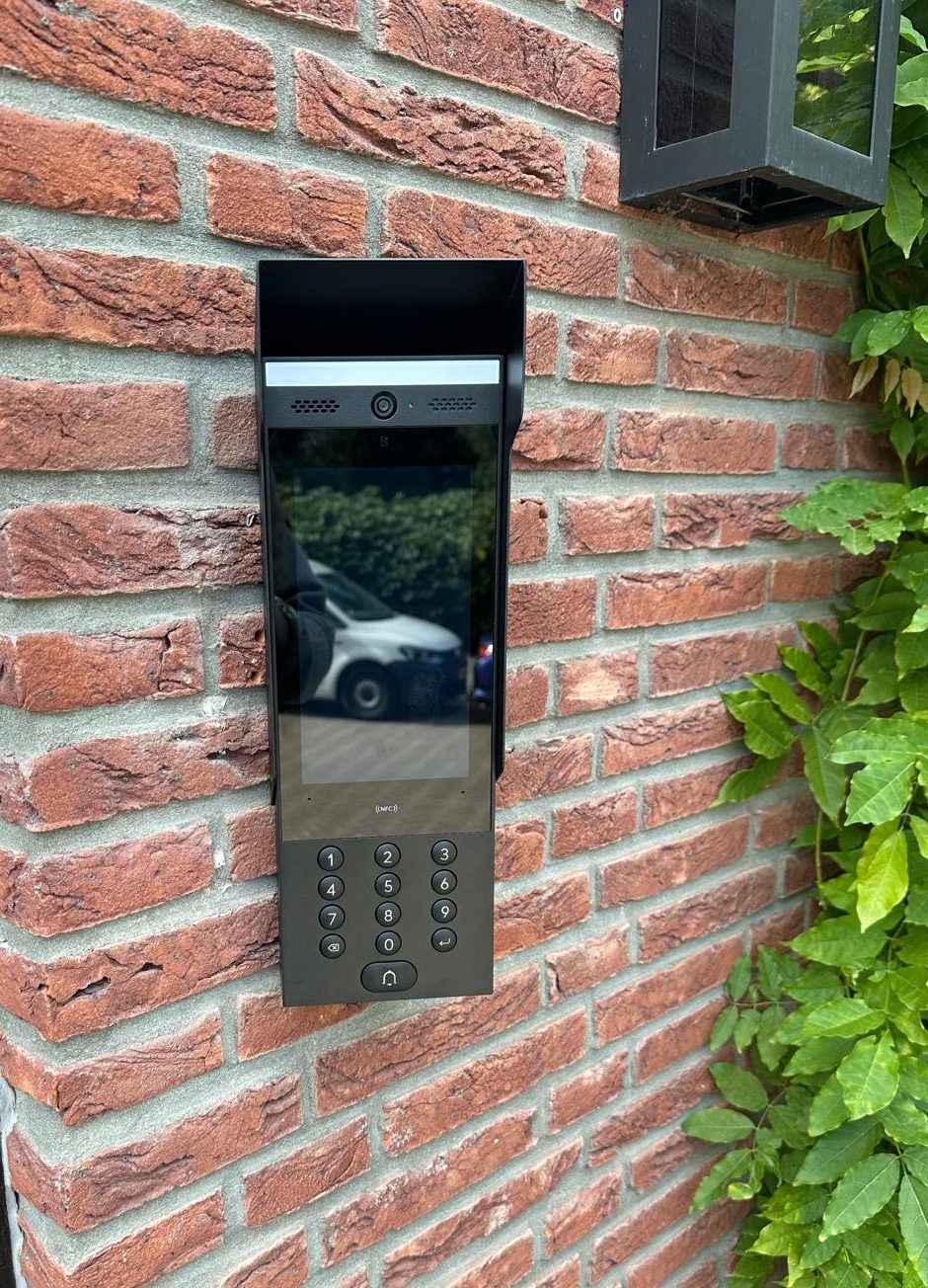 Video-intercom met scherm en codeklavier op een bakstenen buitenmuur, met daarboven een buitenlamp en klimop langs de gevel