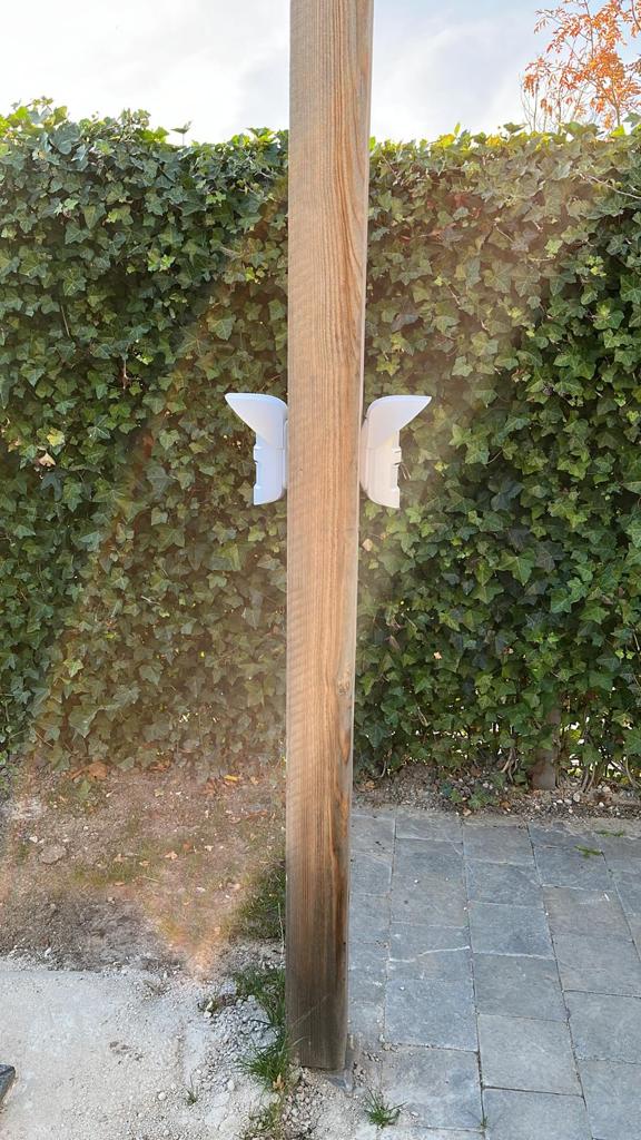 Twee witte buitensensoren gemonteerd op een houten paal voor een groene klimop-haag, om beweging op het terrein te detecteren