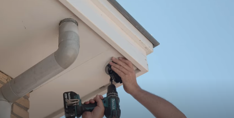 Monteur schroeft met een accuboormachine een IP-camera vast onder de witte dakrand van een woning