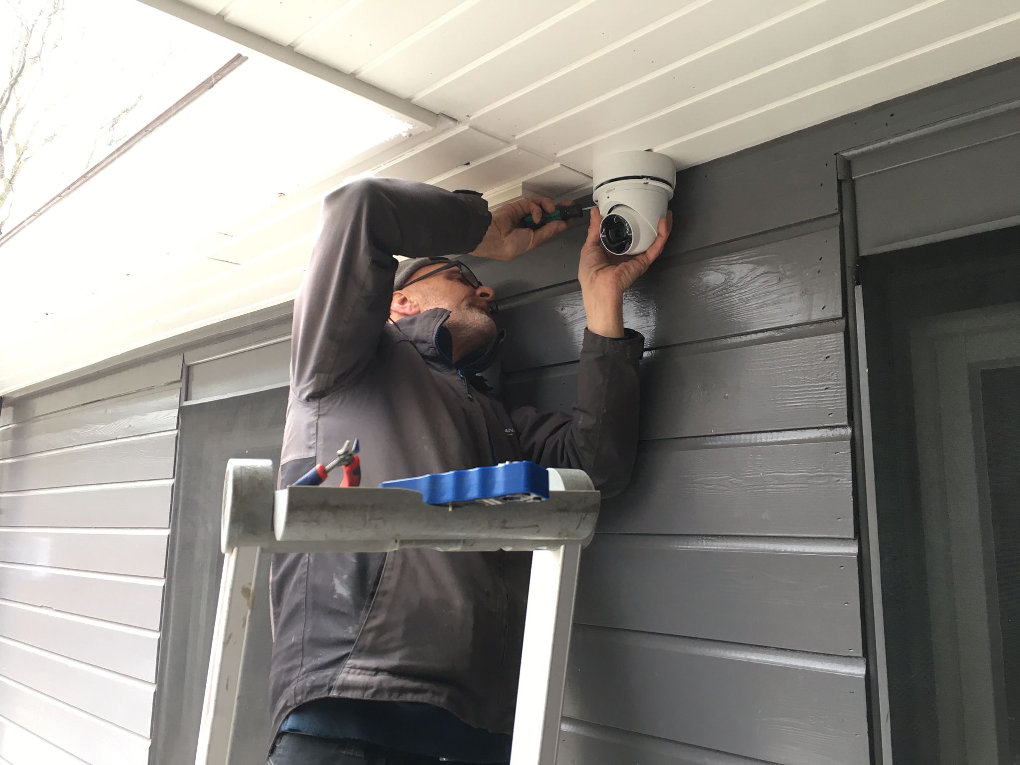 Monteur van CameraInstallatie schroeft een witte dome-camera vast onder de dakrand bij een ingang