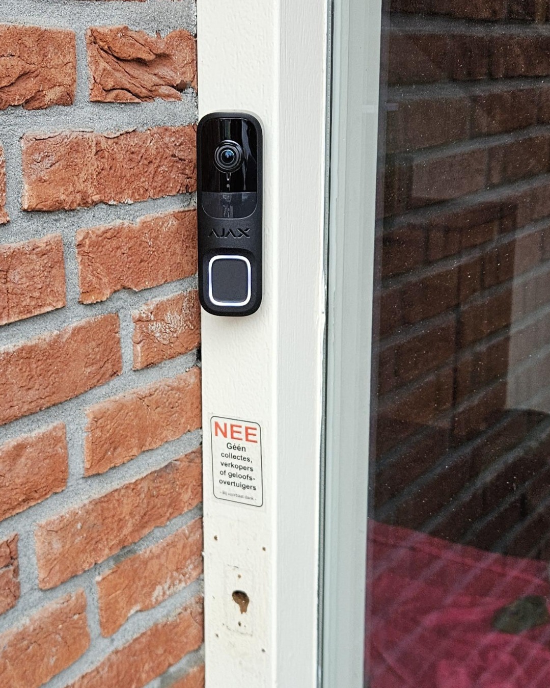 Zwarte Ajax DoorBell gemonteerd op een witte deurstijl naast rode baksteen, ons eigen werk