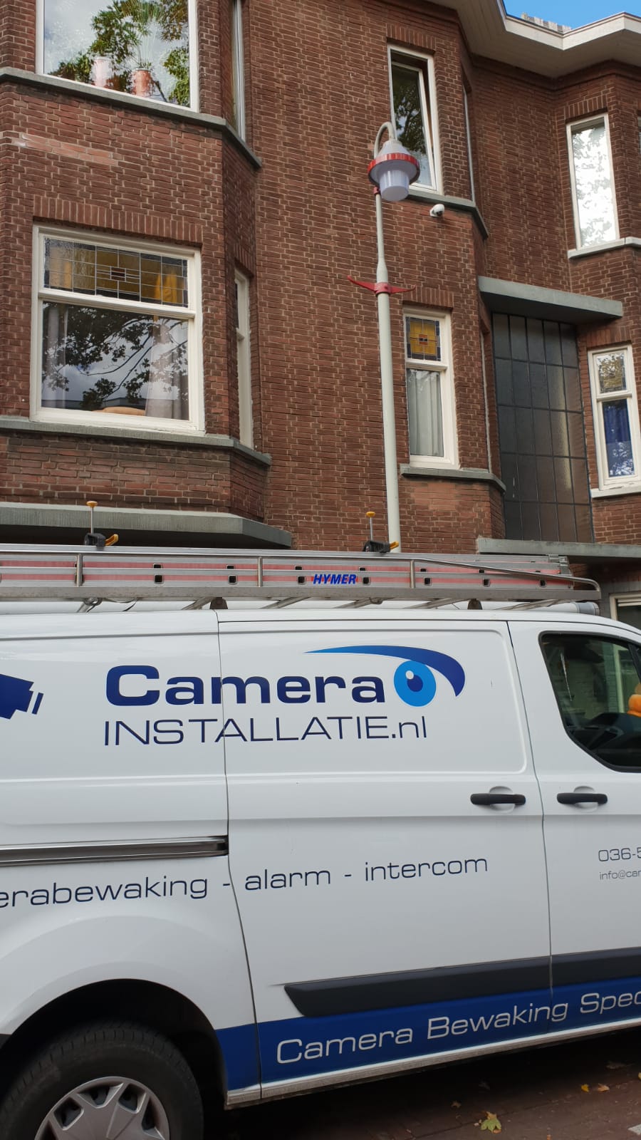 Servicebus van CameraInstallatie geparkeerd voor een pand, met de ladder op het dak