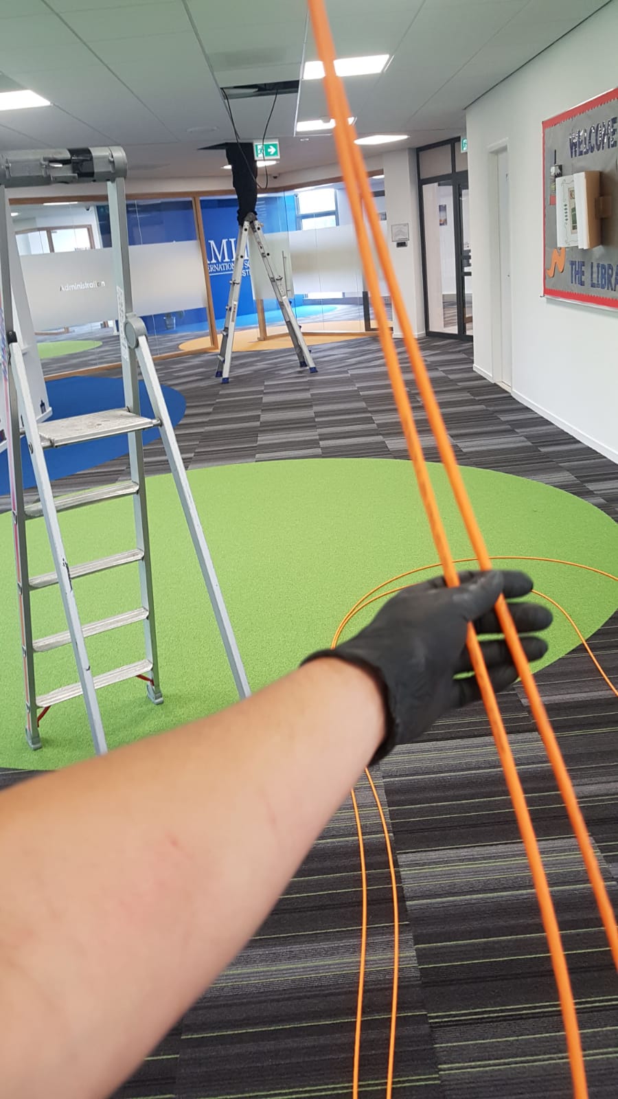 Monteur trekt met de hand een oranje netwerkkabel door een gang, met de ladder verderop