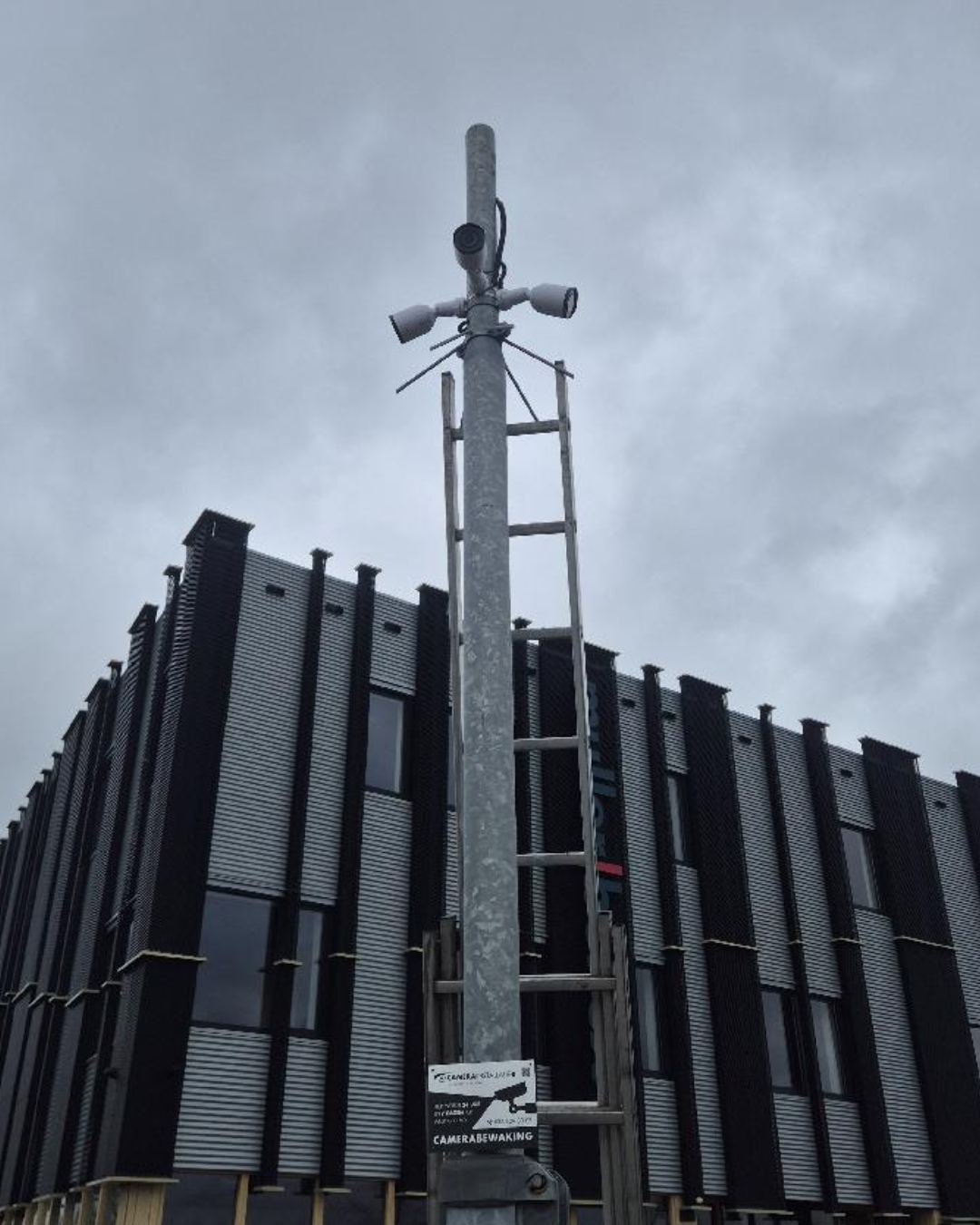 Verzinkte mast met drie camera&rsquo;s bovenin, een ladder ertegenaan en het bord camerabewaking eronder