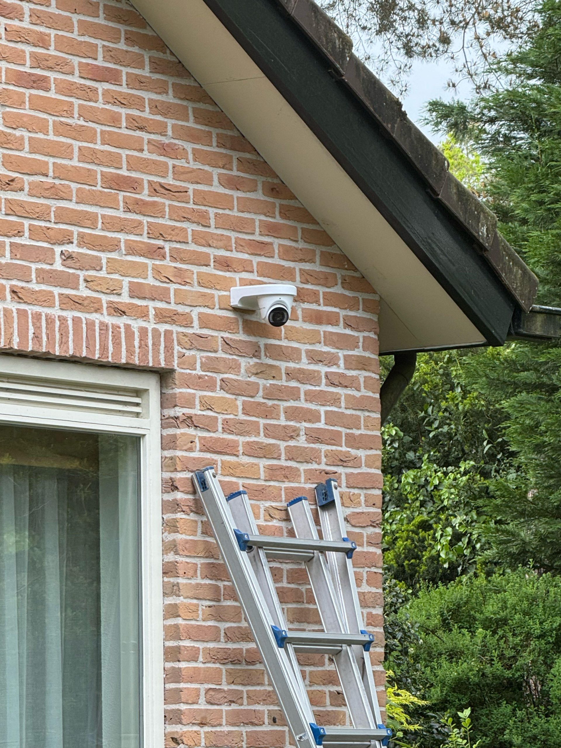 Witte camera onder de dakrand van een woning, met de ladder er nog tegenaan