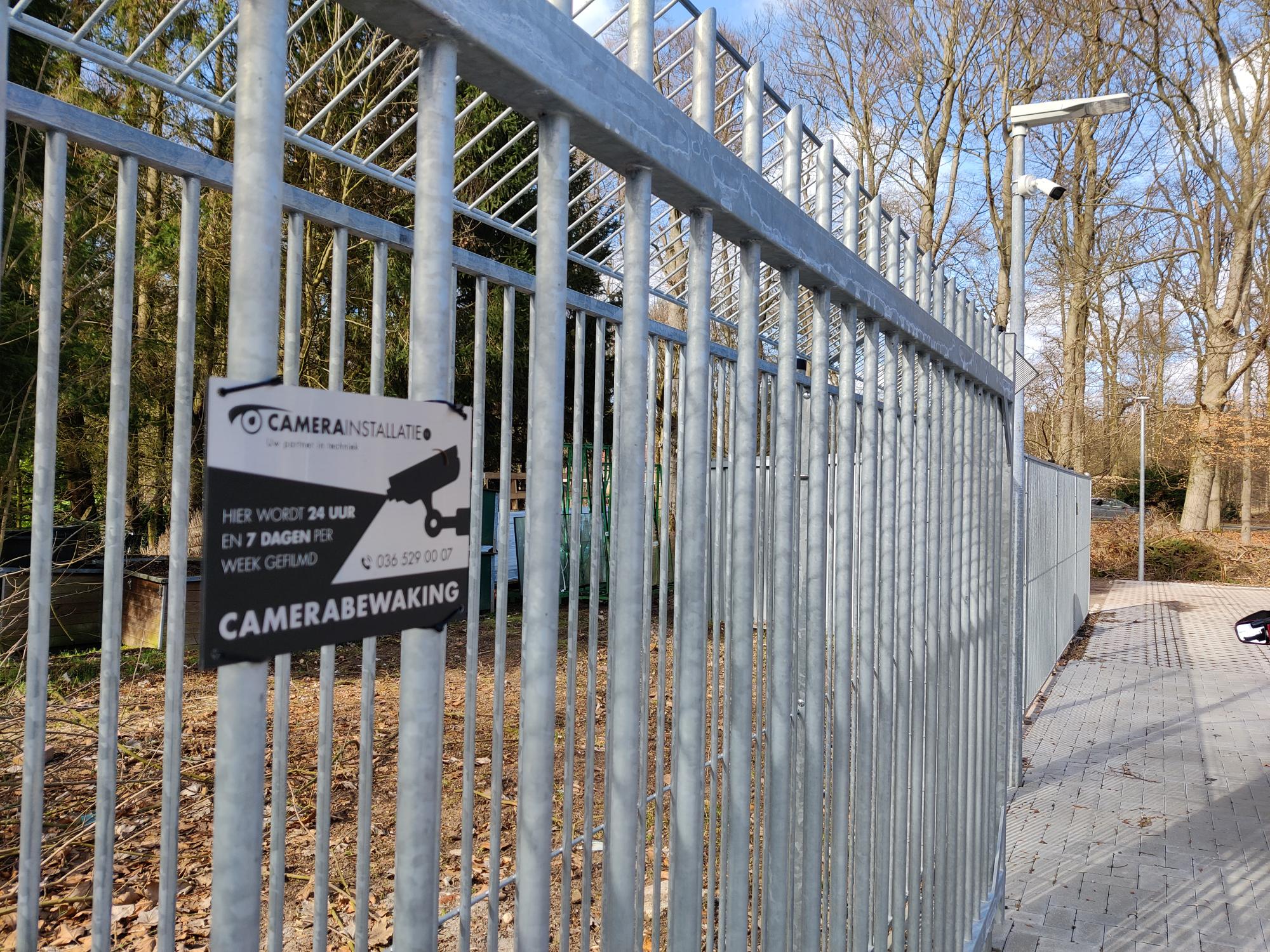 Bordje met de tekst camerabewaking aan een hek, met een beveiligingscamera erachter
