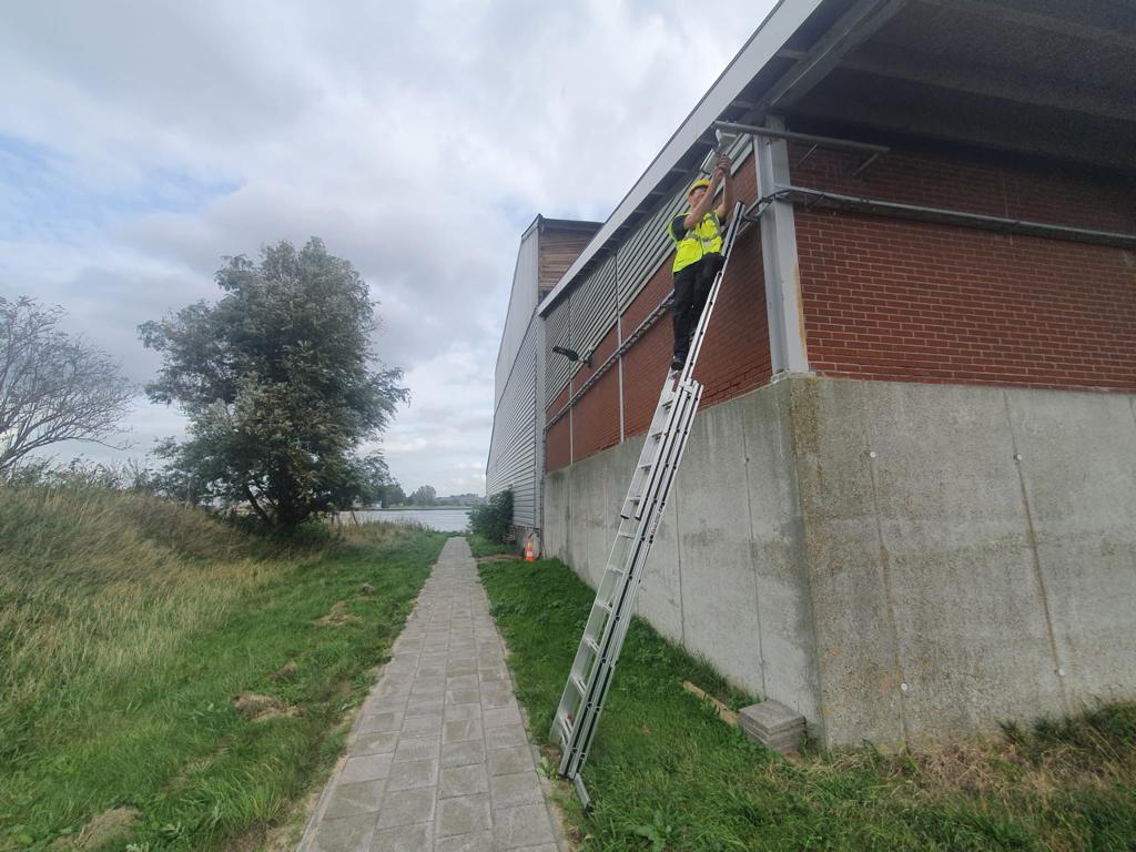 Monteur in geel hesje op een ladder monteert een camera onder de dakrand van een grote loods aan het water