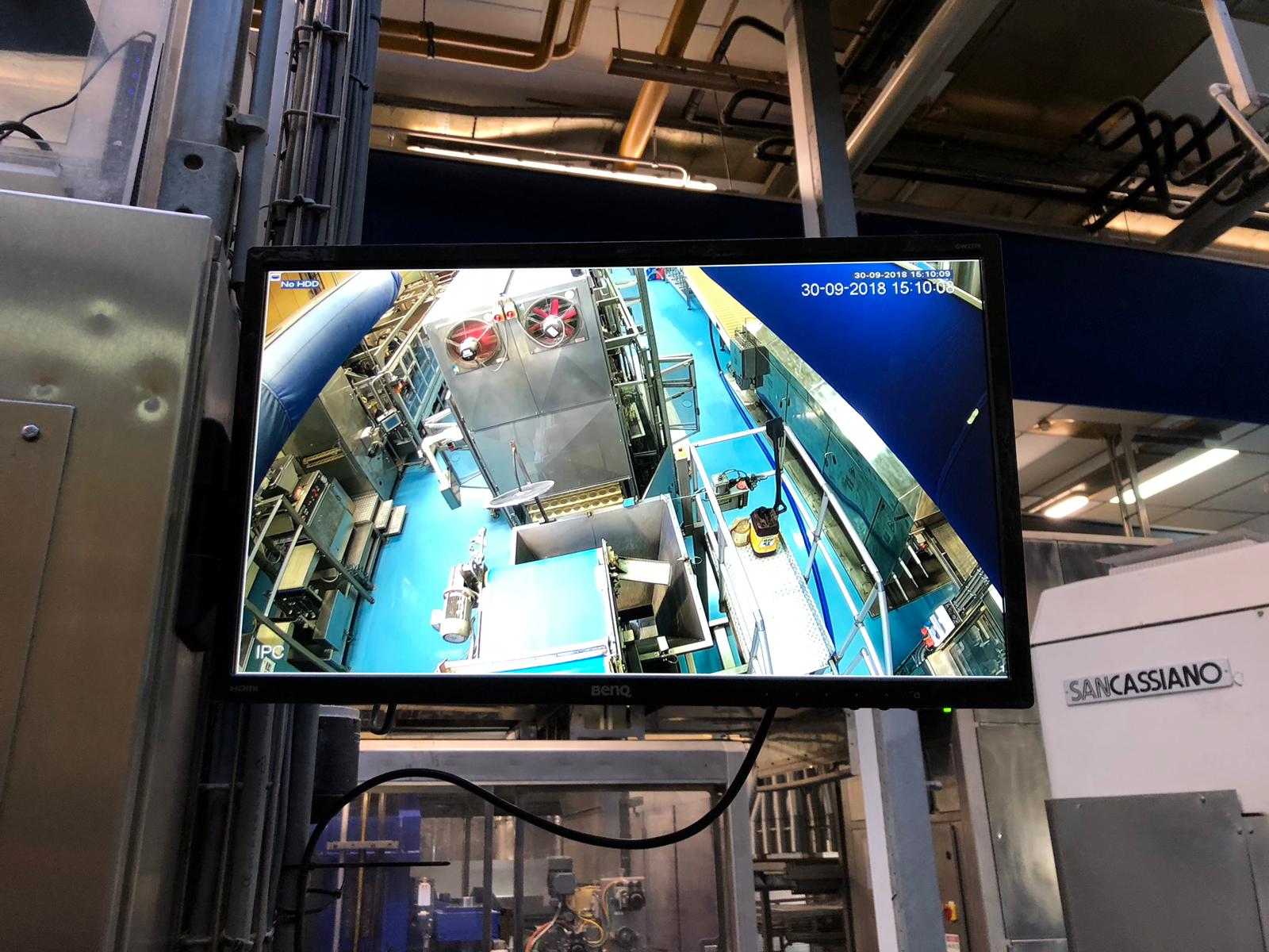 Scherm aan een stalen constructie in een productiehal toont live camerabeeld van de werkvloer met roestvrijstalen machines