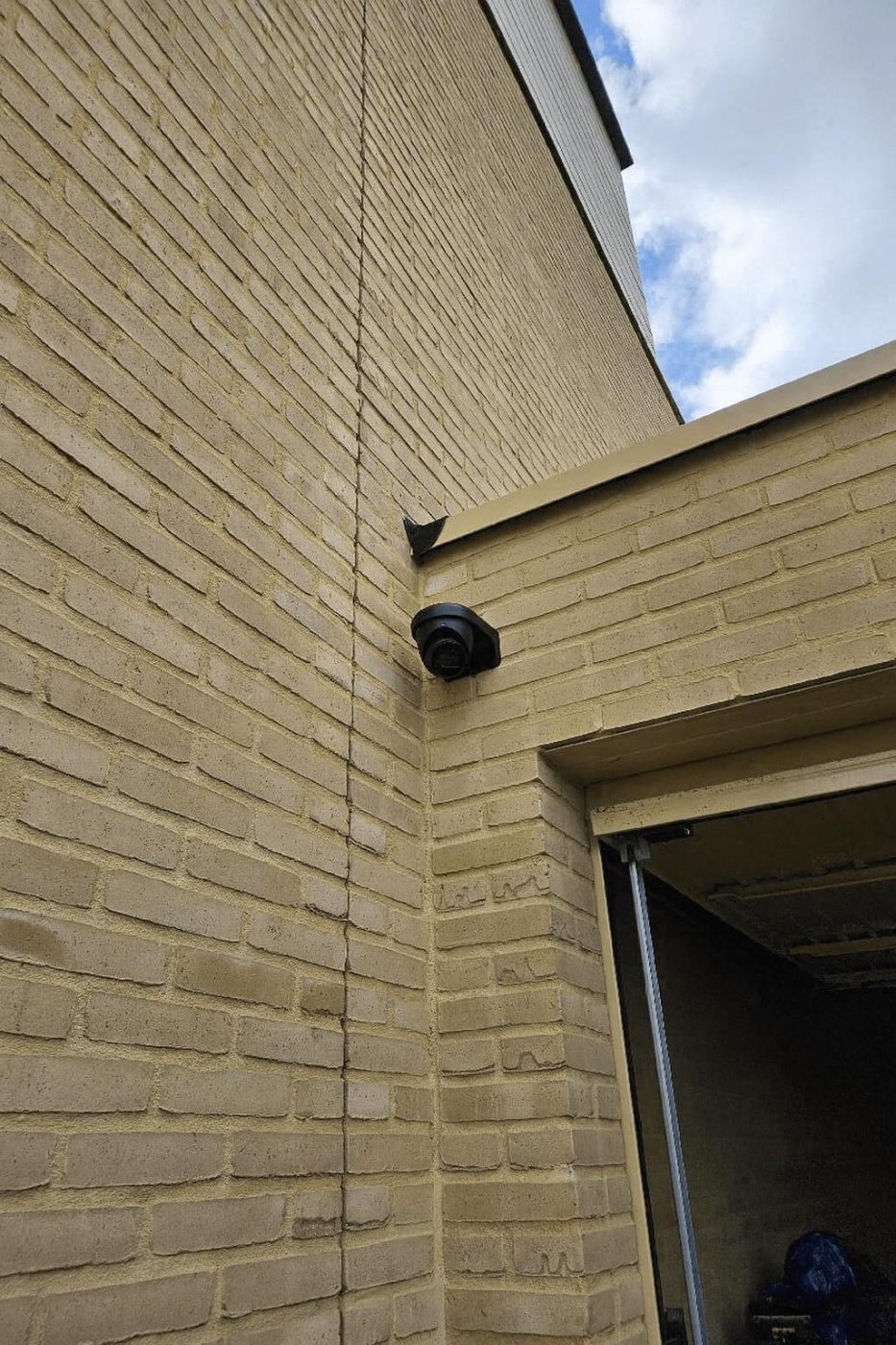 Zwarte dome-camera gemonteerd in een buitenhoek van een lichtgekleurde bakstenen gevel bij de ingang van een bijgebouw