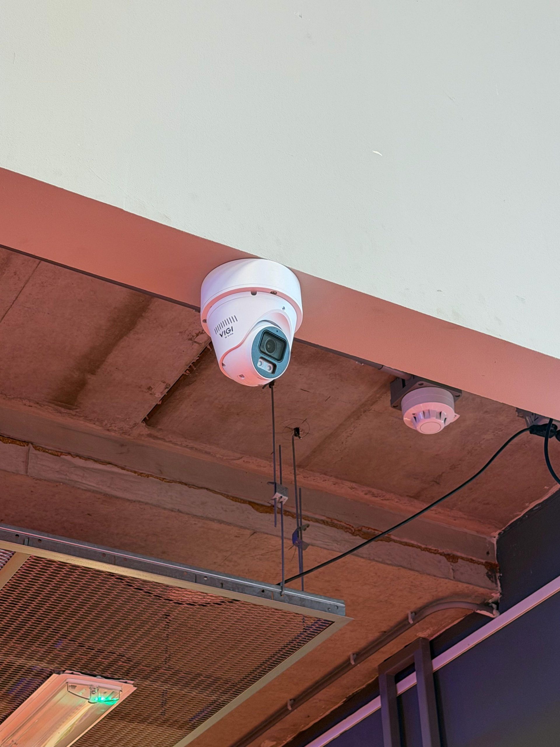 Witte VIGI-camera onder een roodgeschilderde balk, naast een rookmelder in een openbare ruimte