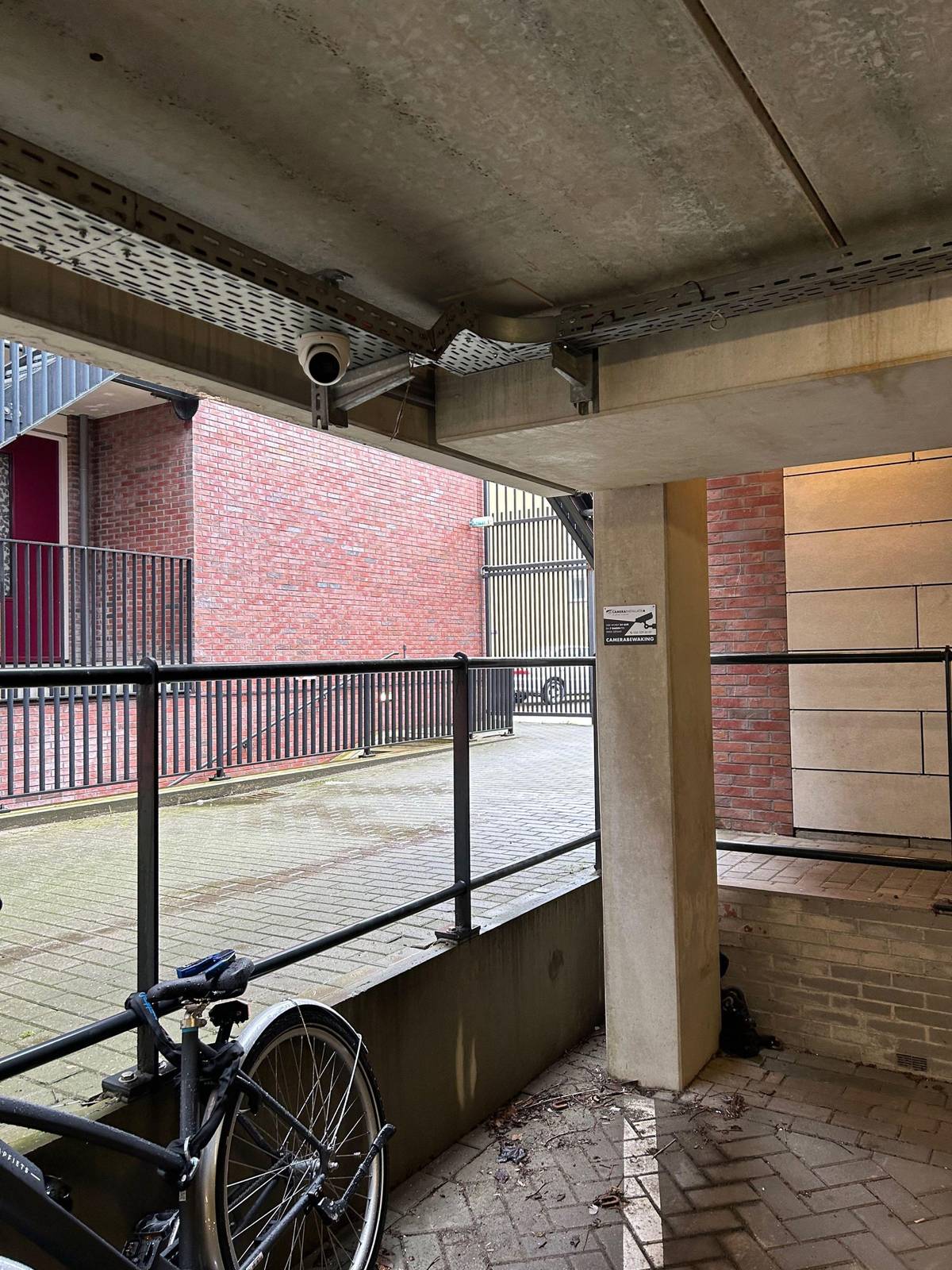 Camera boven een overdekte stalling met fietsen en een scooter, met een sticker camerabewaking op de kolom