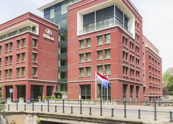 Het hotelpand in Den Haag gezien vanaf de brug over de gracht, met de hoofdentree en de Nederlandse vlag