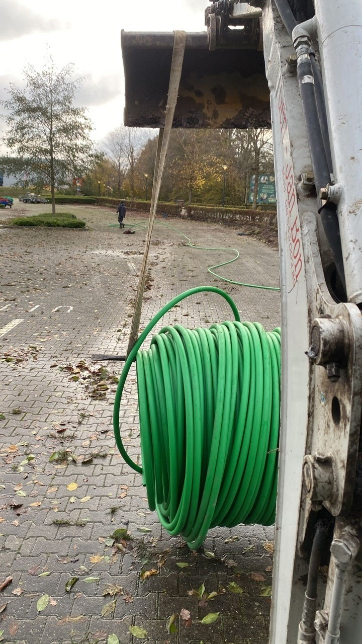 Rol groene mantelbuis aan de arm van een minigraver, terwijl de buis over het terrein wordt uitgelegd