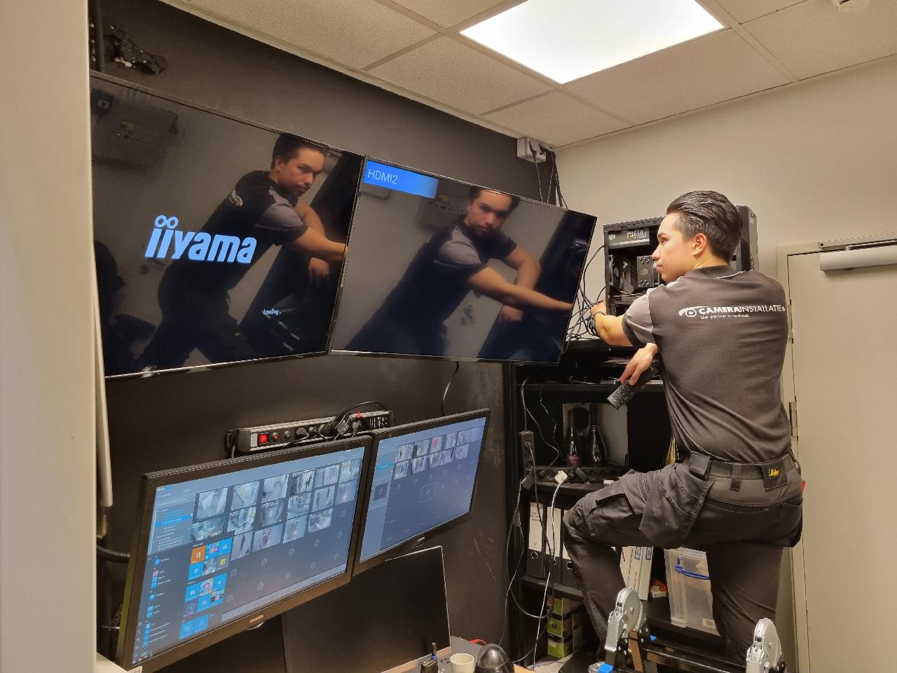 Monteur van CameraInstallatie werkt in een uitkijkruimte aan de cameraserver, met wandschermen die live camerabeelden en een cameraraster tonen