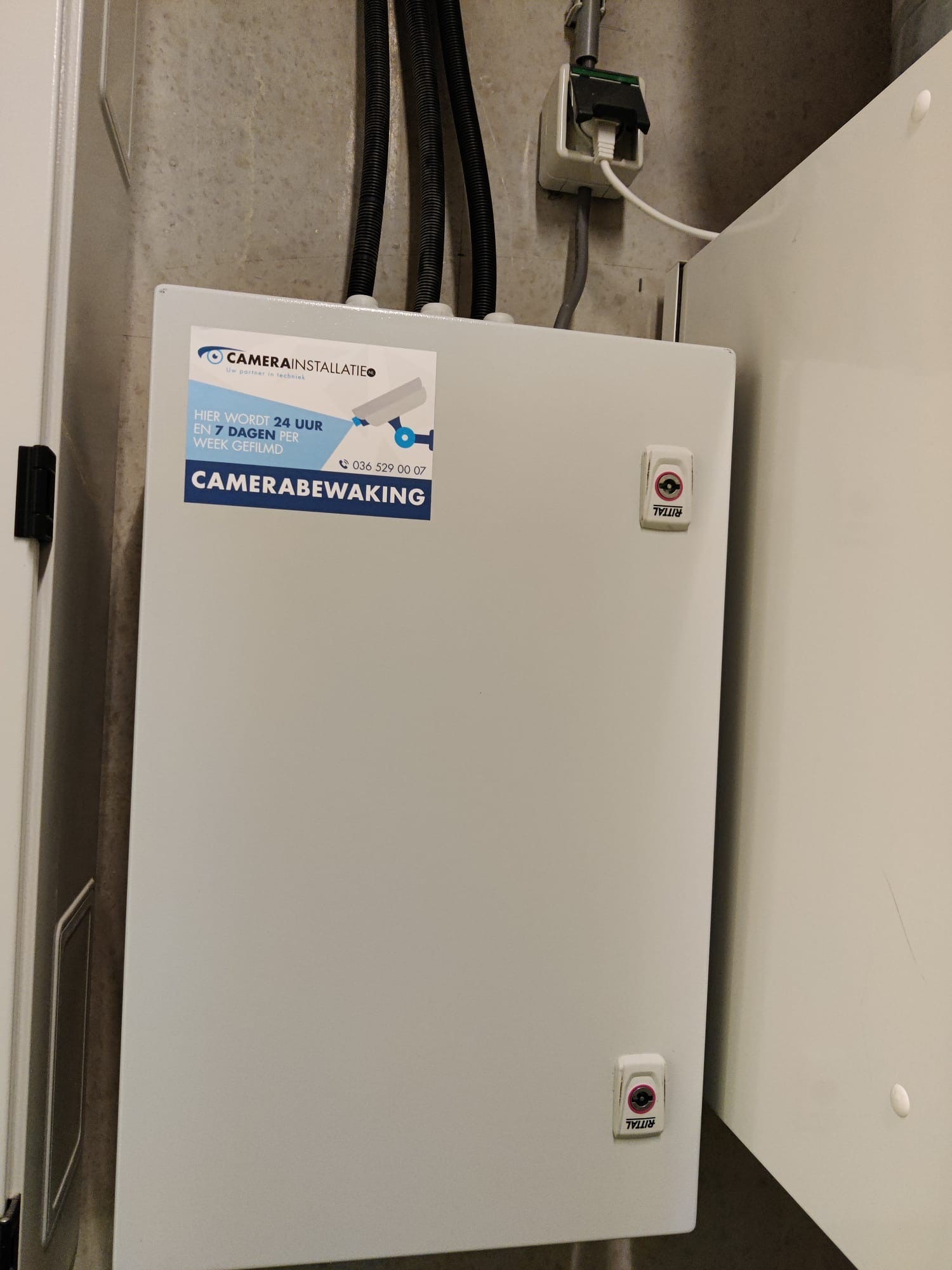 Witte Rittal-netwerkkast met een sticker van CameraInstallatie: hier wordt 24 uur en 7 dagen per week gefilmd
